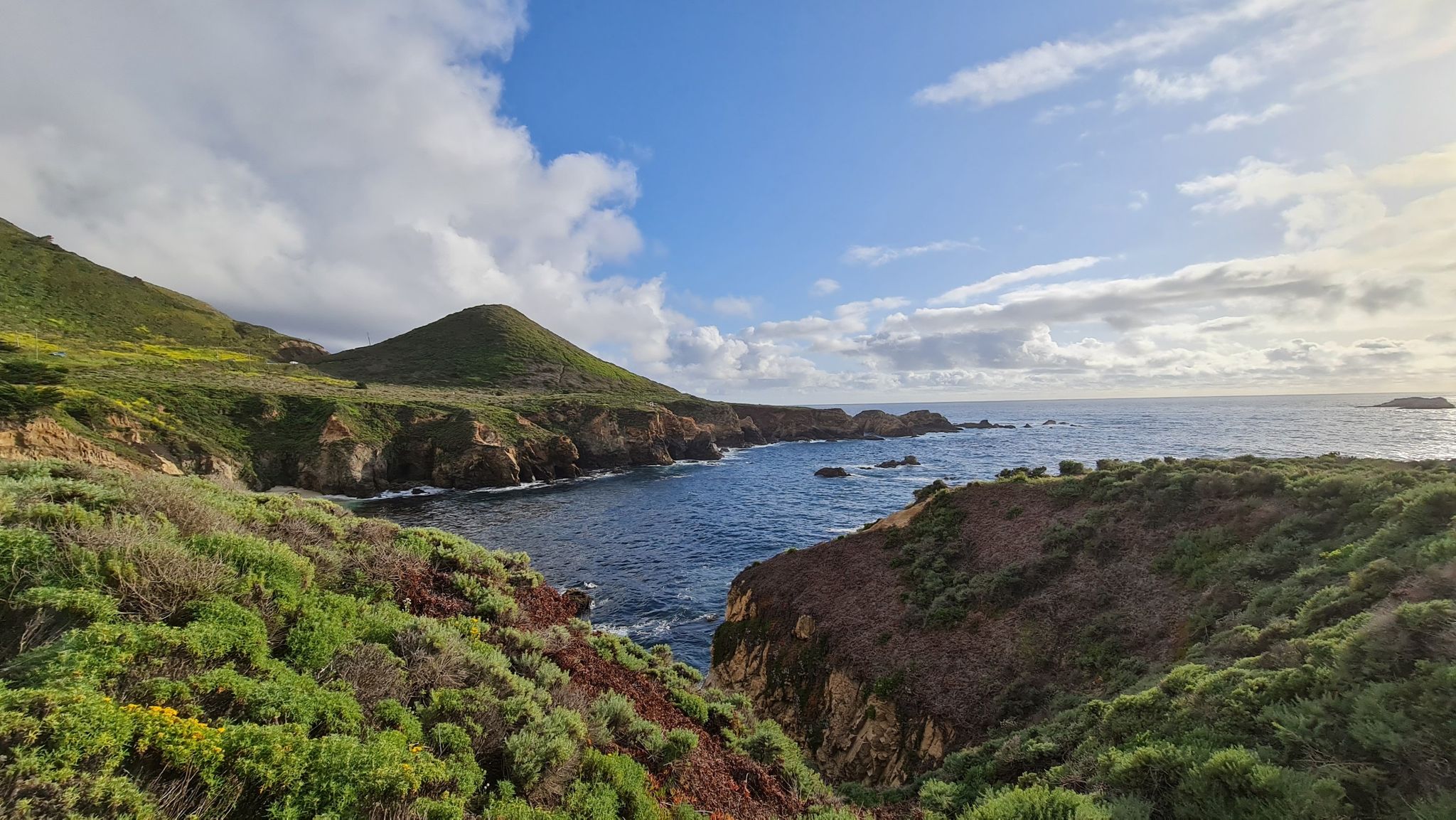Coastal landscape with green hills, rocky cliffs, and blue ocean under a partly cloudy sky. - Garrapata Bluff Trail