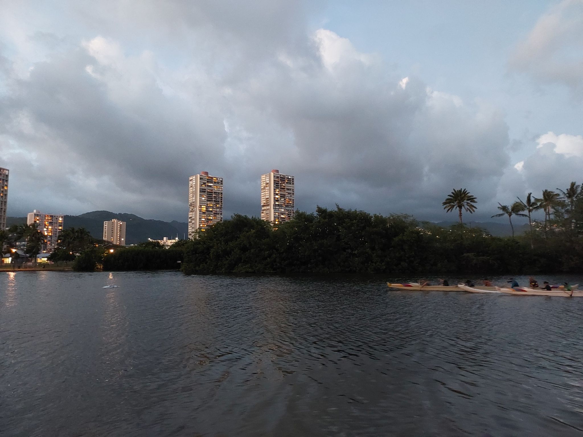 City skyline with tall buildings, calm water, and a rowboat at dusk. - Ala Wai Park Trail