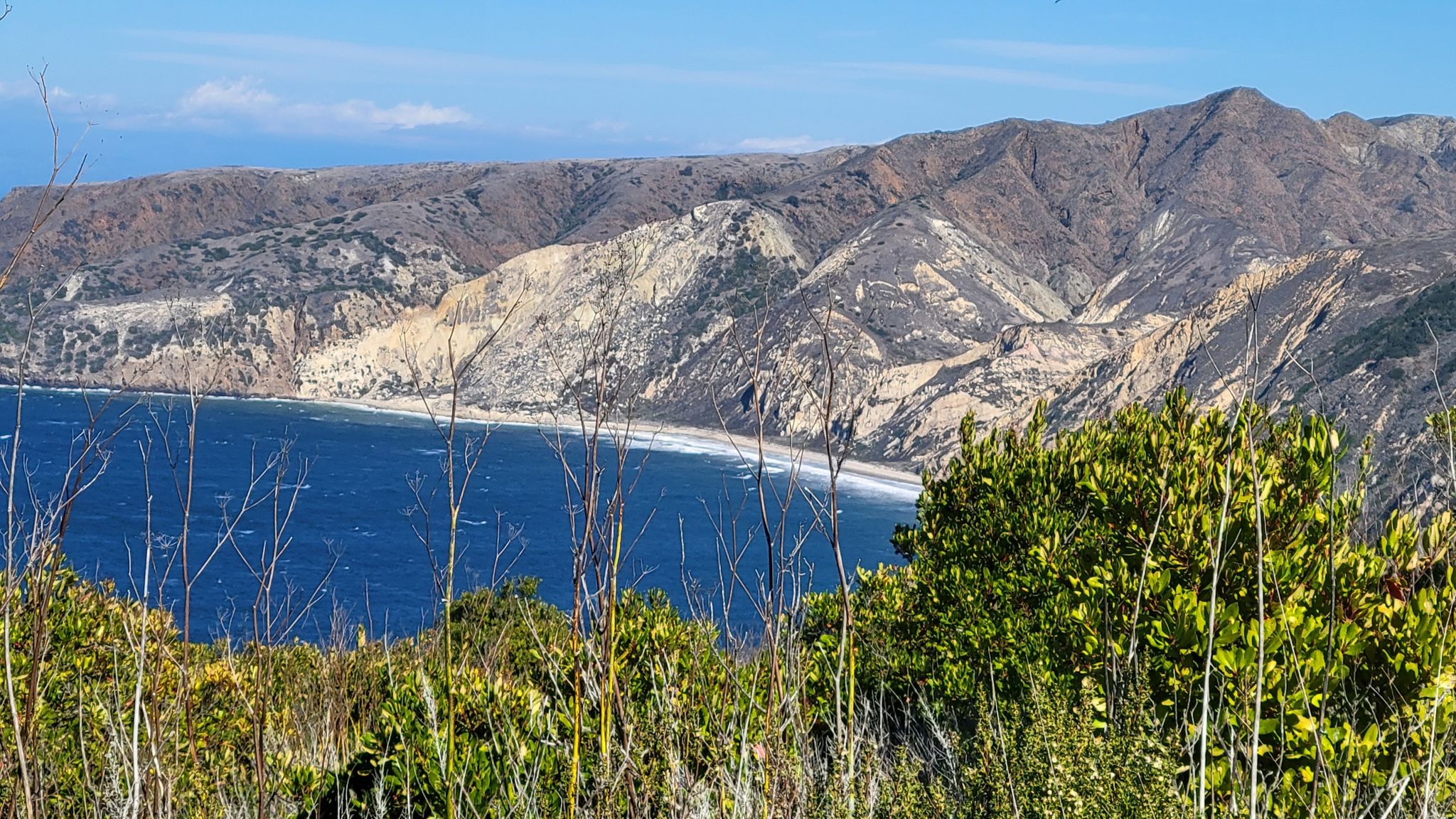Bright coastal view of blue ocean, sandy beach, and rugged multi-colored cliffs. - Montañon Ridge Loop Trail