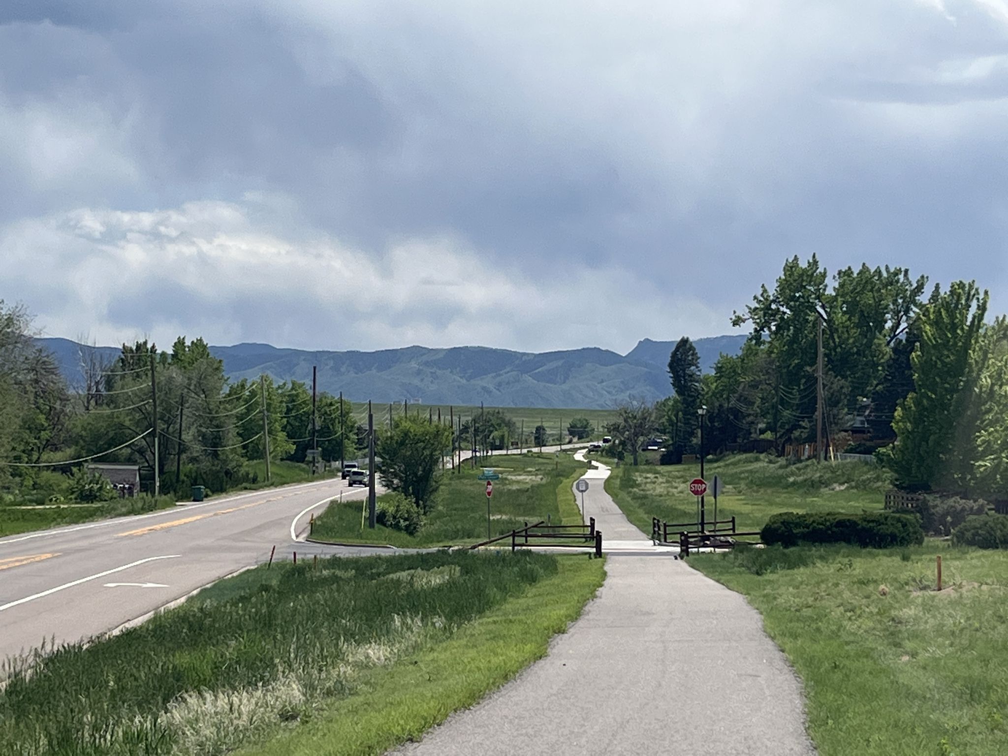 A paved road and path run parallel towards distant mountains under a cloudy sky. - Columbine Trail