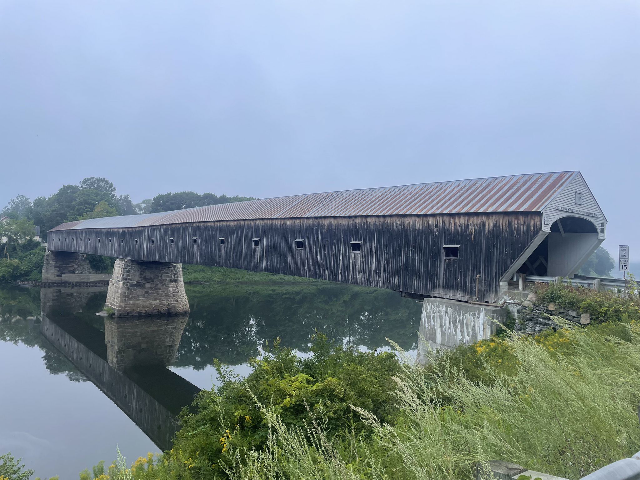 Long wooden covered bridge spans a calm river under an overcast sky. - Plainfield loop from Cornish