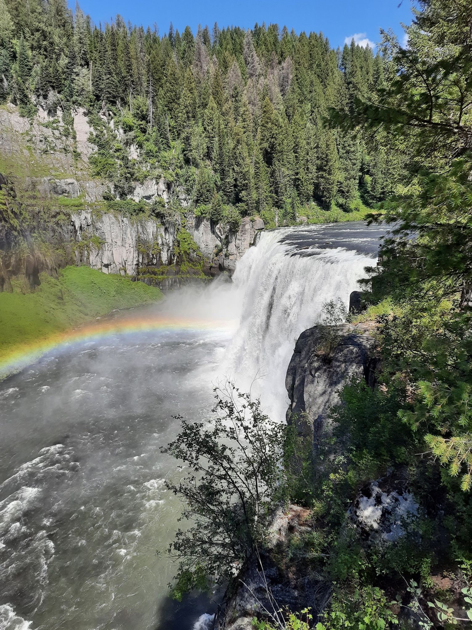 A powerful waterfall plunges into a river, with a rainbow in the mist. - Upper Mesa Falls Boardwalk