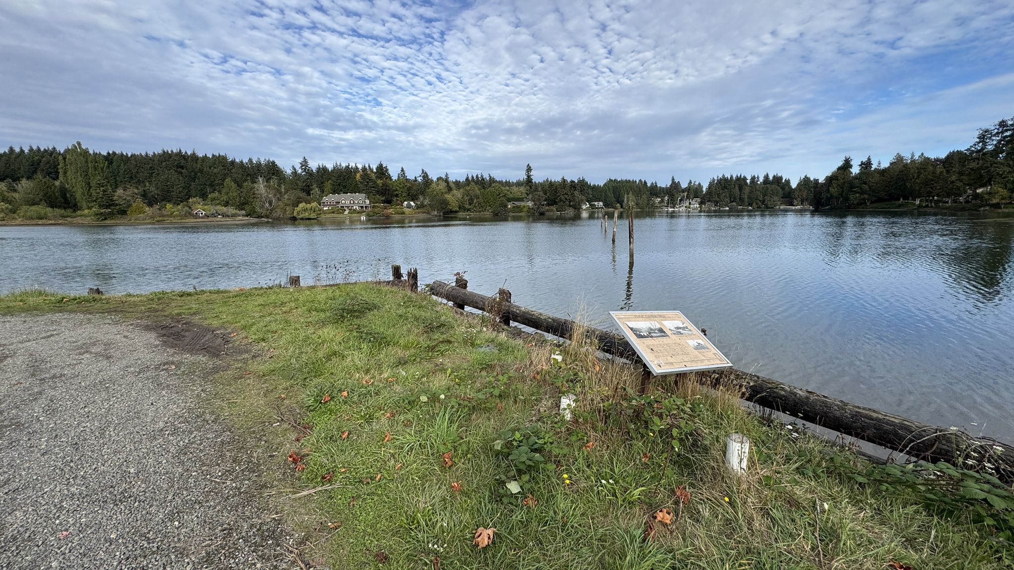Wide river with a grassy bank, sign, and forested shore under cloudy sky. - Hall Hill Labyrinth – View of Blakely Harbor loop from Fort Ward Park