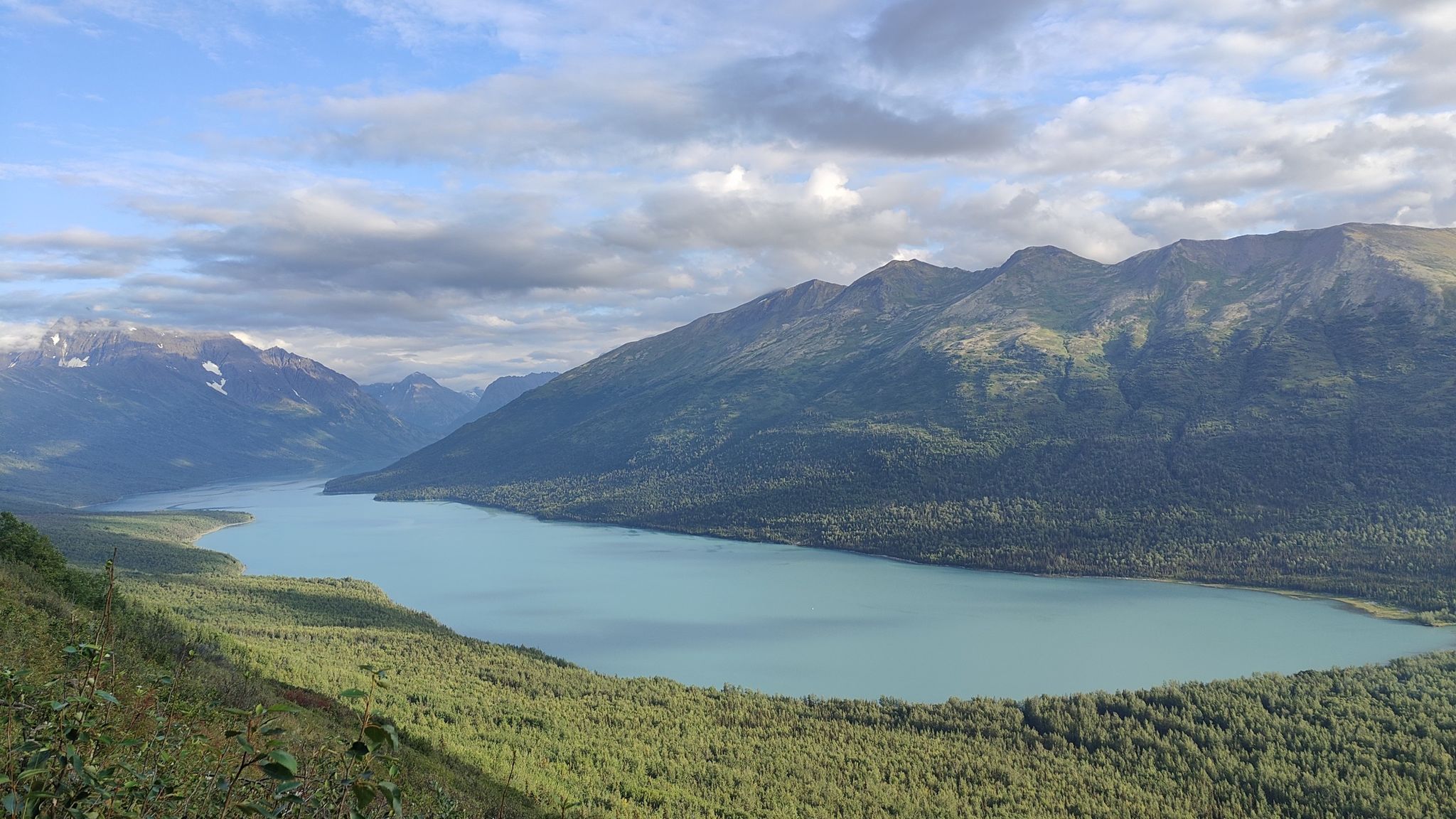 Wide view of a large, calm lake surrounded by forested mountains under cloudy sky. - Twin Peaks Trail