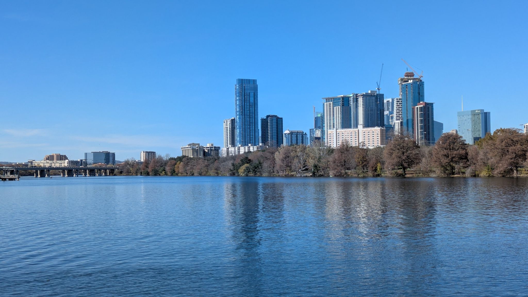 City skyline across a wide river with a distant bridge under a clear sky. - Boardwalk – Longhorn Point loop from East Riverside - Oltorf