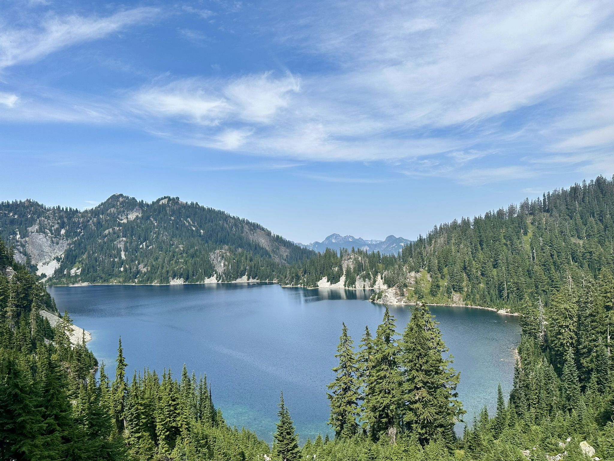 A clear blue lake nestled among green forested mountains under a bright sky. - Snow Lake Trail