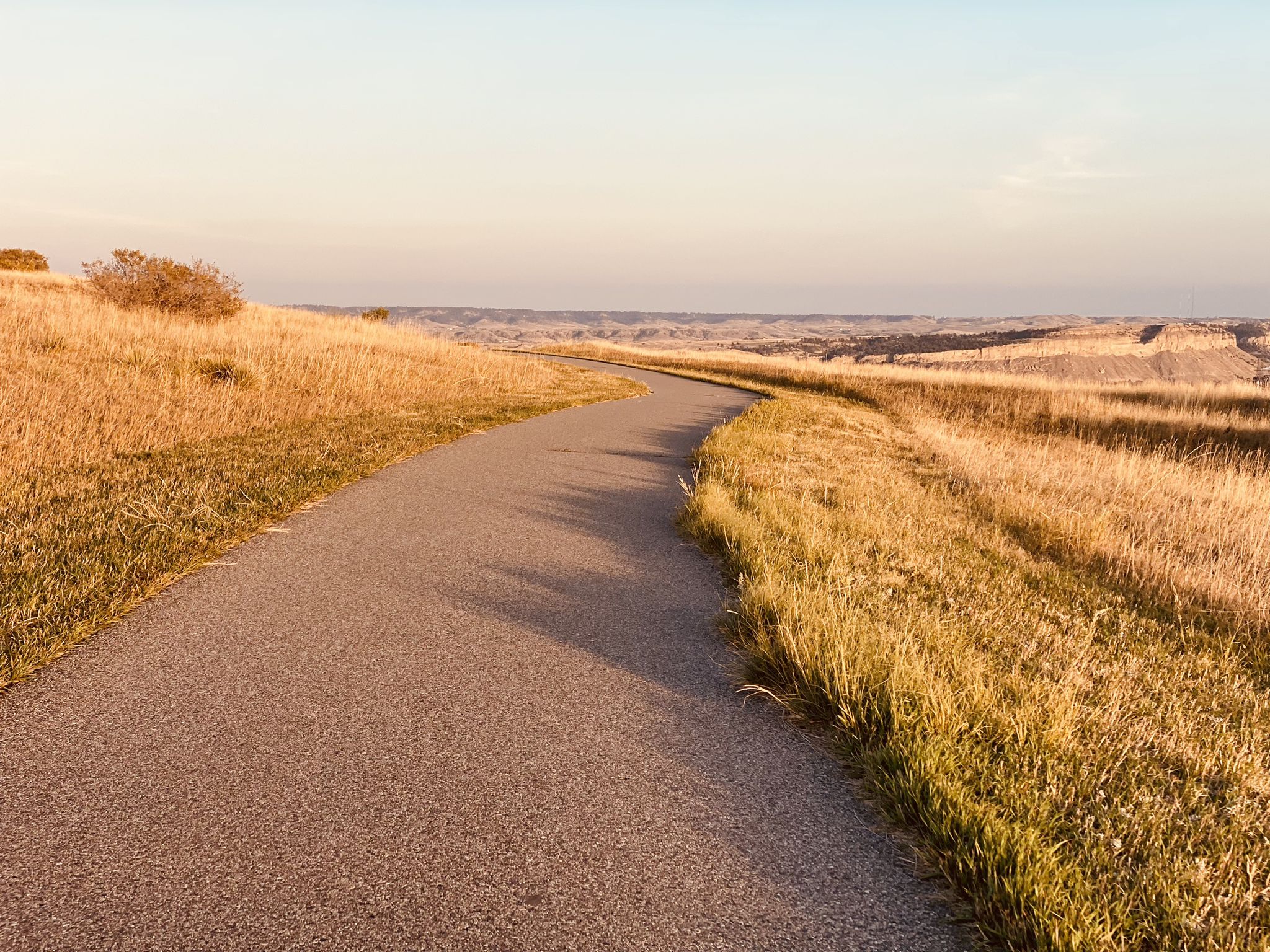 Paved path curving through sunlit golden grass and rolling hills. - Chief Black Otter Trail