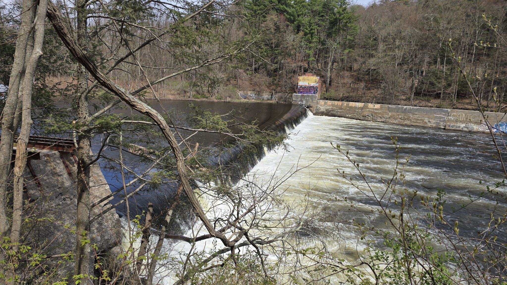Rushing water over a dam with trees and a building in the background. - View of the Farmington River – Farmington River crossing loop from Town of Avon Open Space