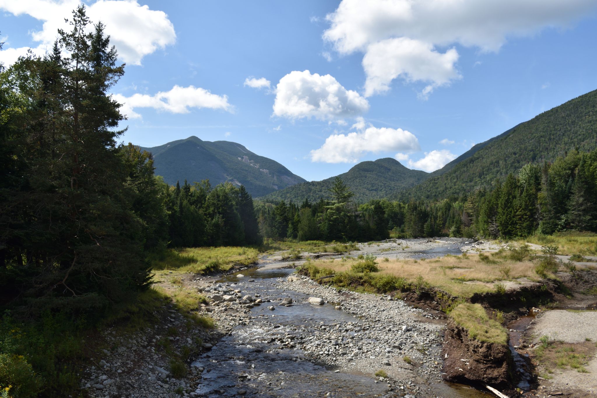A wide, rocky riverbed winding through a valley with forested mountains and clouds. - Mount Marcy via Van Hoevenberg Trail
