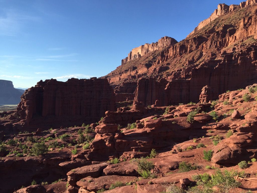 Sunny view of layered red rock cliffs and formations with scattered green shrubs. - Fisher Towers Trailhead loop from Fisher Towers Trailhead