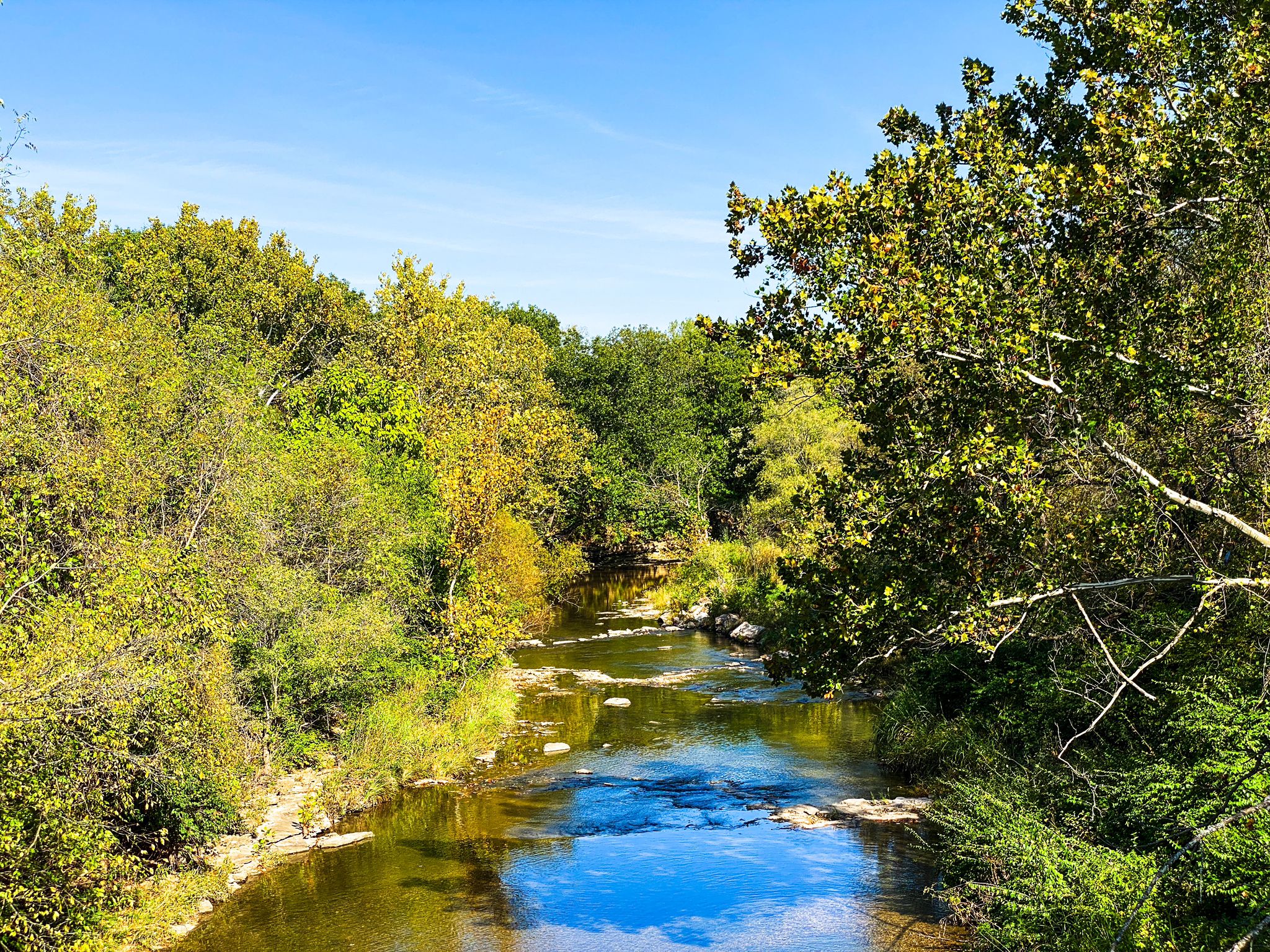 A winding river flows through lush green and yellow trees under a bright blue sky. - Gary L. Haller Trail – Pump Track loop from Lake Quivira