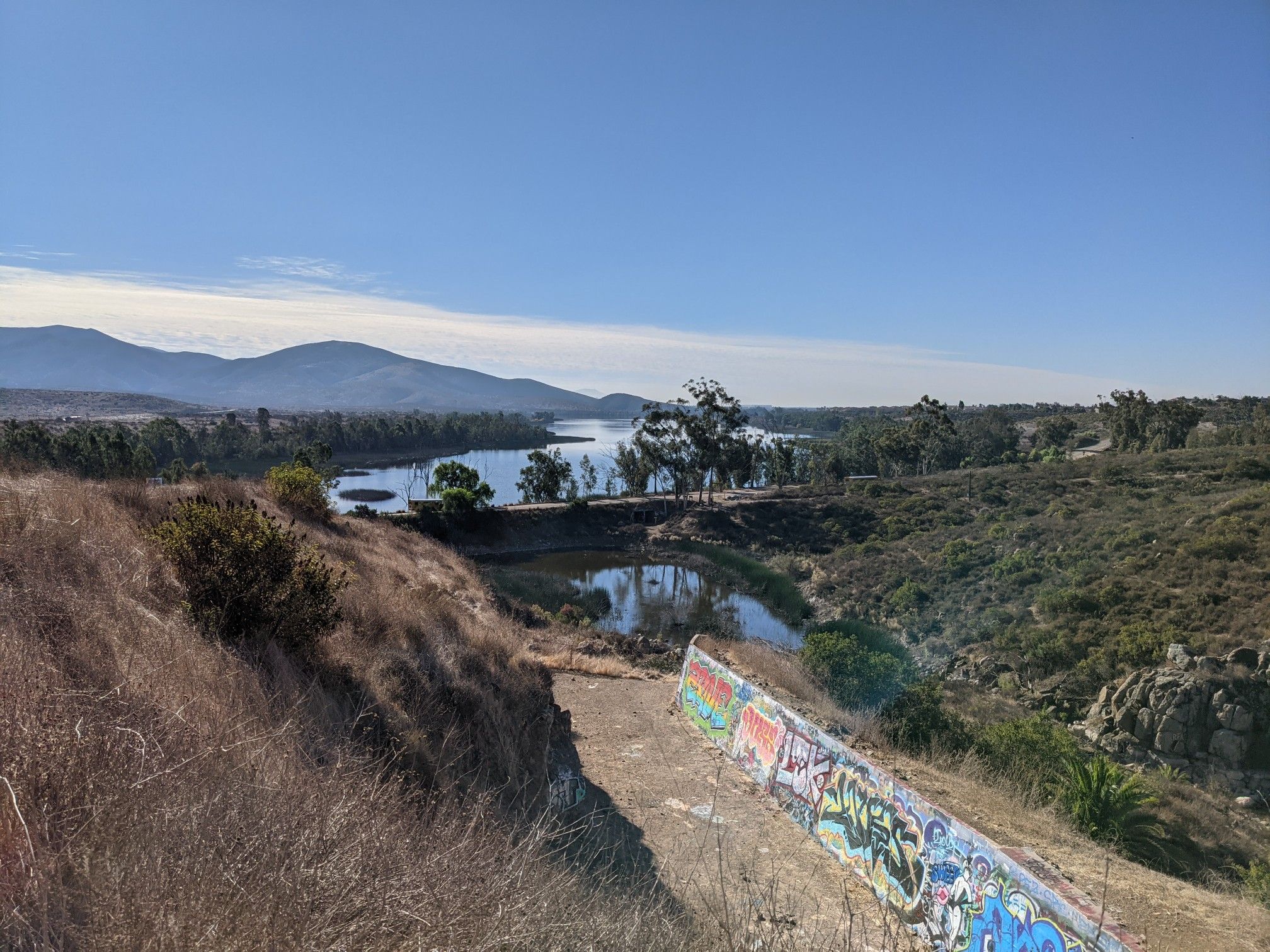 Expansive lake and mountains, with a graffiti-covered wall in the foreground. - Upper Otay Reservoir – Lower Otay Reservoir loop from Otay Ranch Town Center