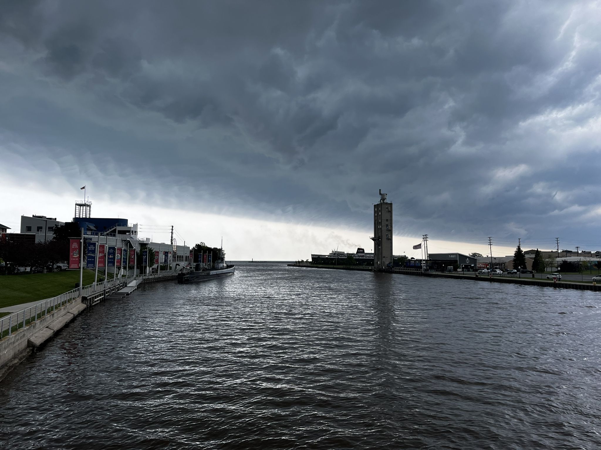 Dark, stormy clouds over a wide river with buildings and a tall tower. - Mariners Trail