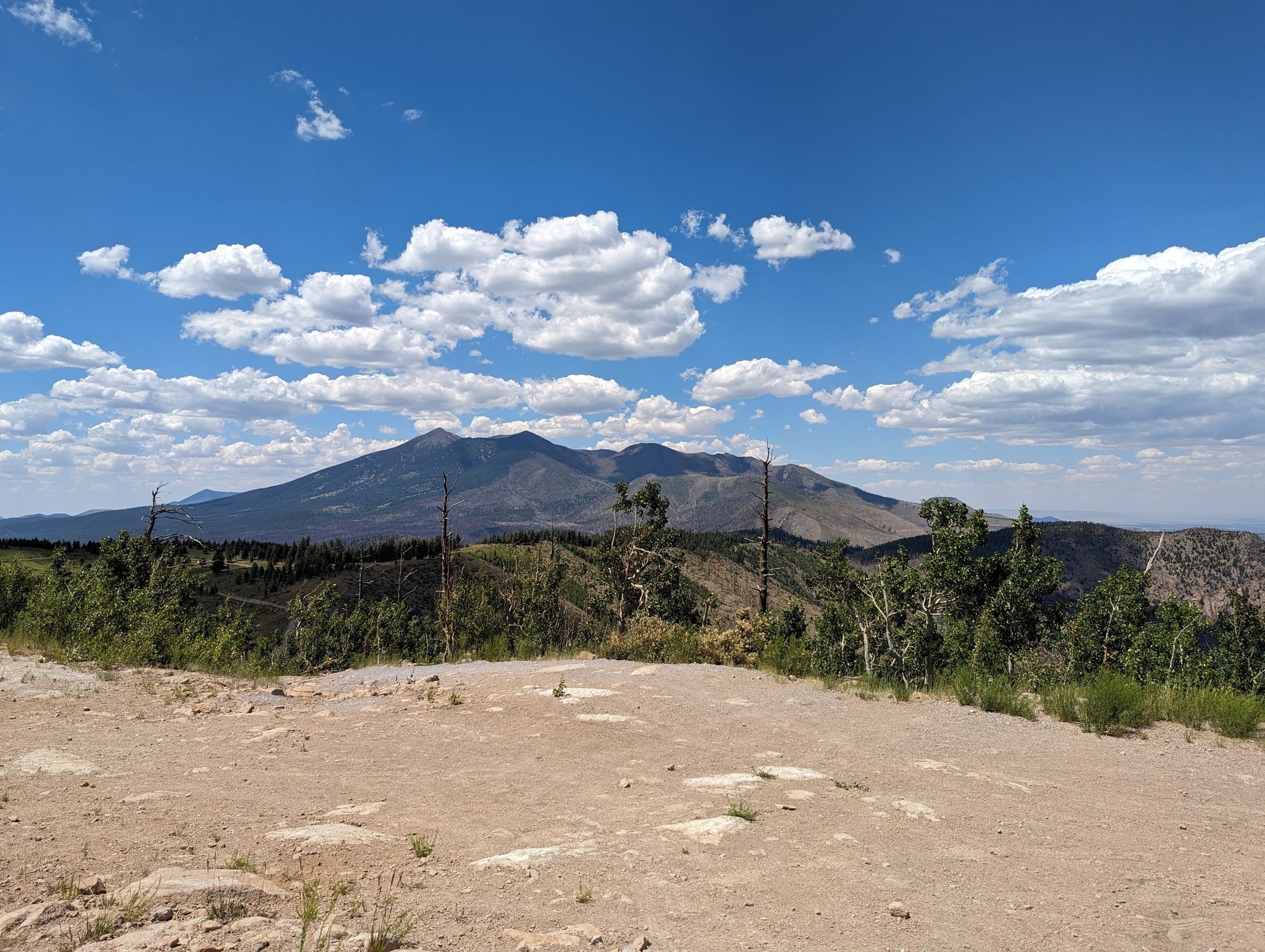 A wide dirt clearing overlooks a vast mountain range under a bright, cloudy sky. - Sunset Trail