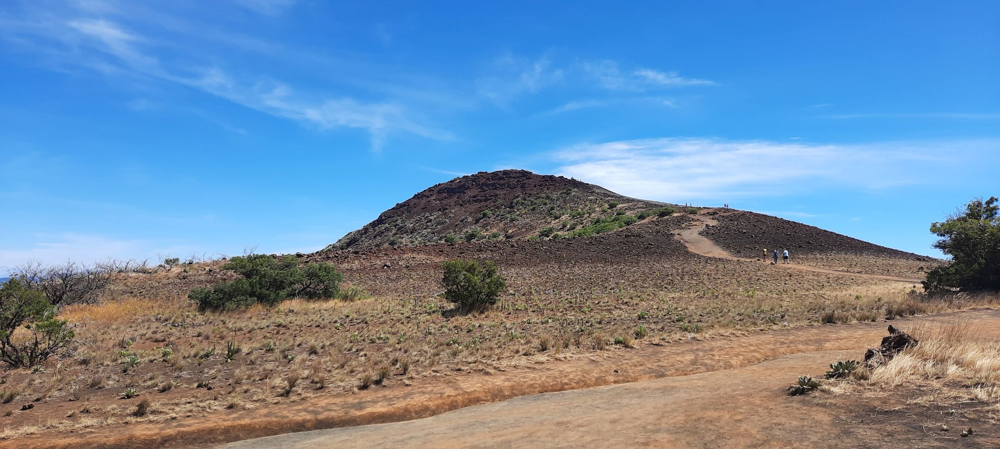 Sunny view of a dark, dry hill with a winding path and distant hikers. - Pu‘ukalepeamoa Lookout