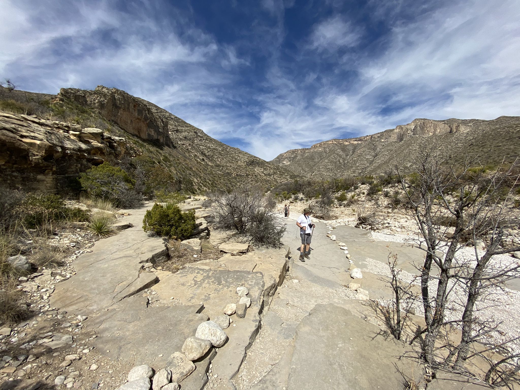 A person hikes a rocky desert path between large mountains under a bright sky. - McKittrick Canyon Trail