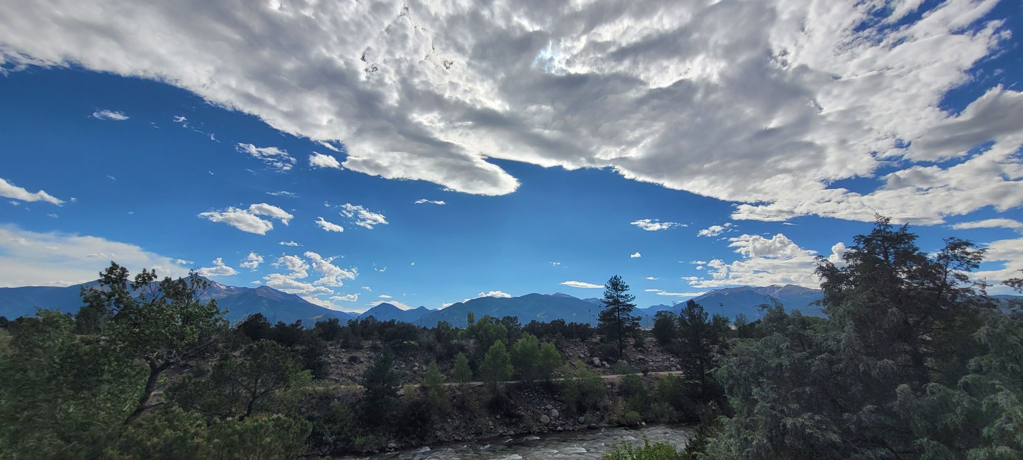 A river flows through a forested landscape with distant mountains under a dramatic sky. - Bridge to Bridge Trail