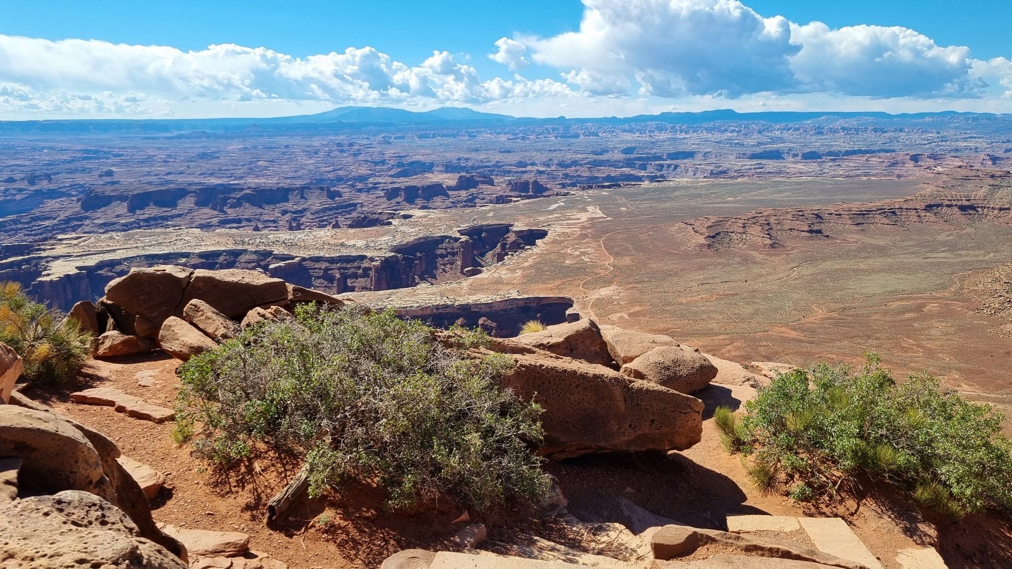 Rocky outcrop with green bushes overlooking a vast canyon under a partly cloudy sky. - Grand View Point Trail