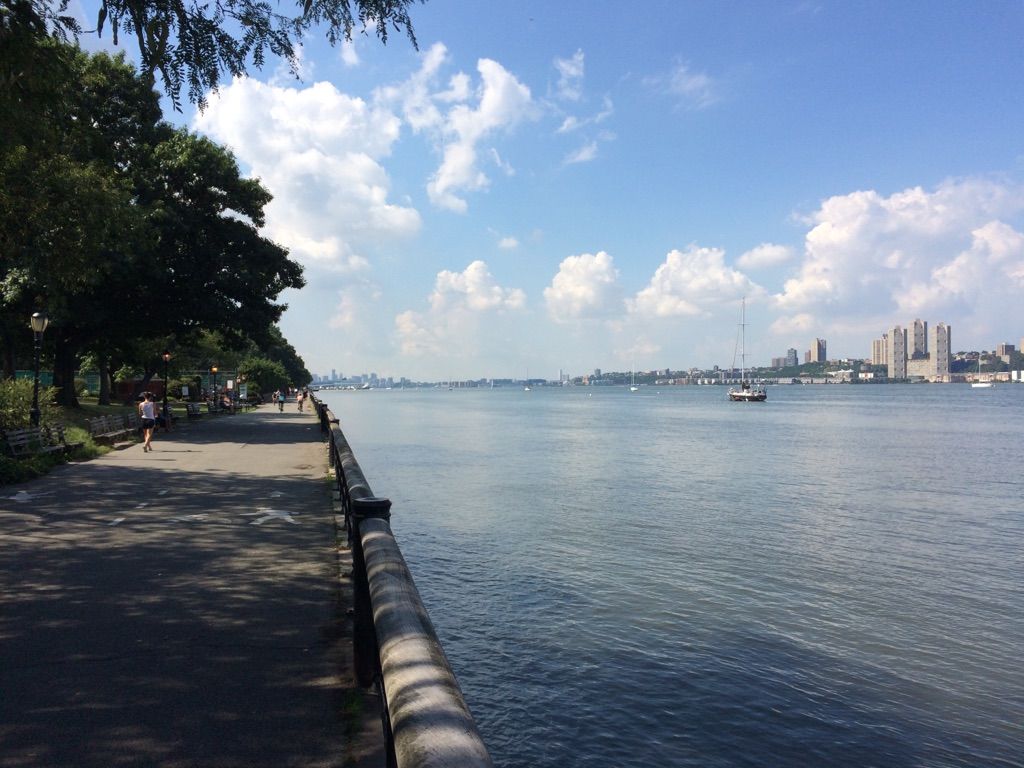 Paved path along a river with people walking, distant city, and boats. - Hudson River Greenway – Hudson River Greenway loop from 116th Street–Columbia University