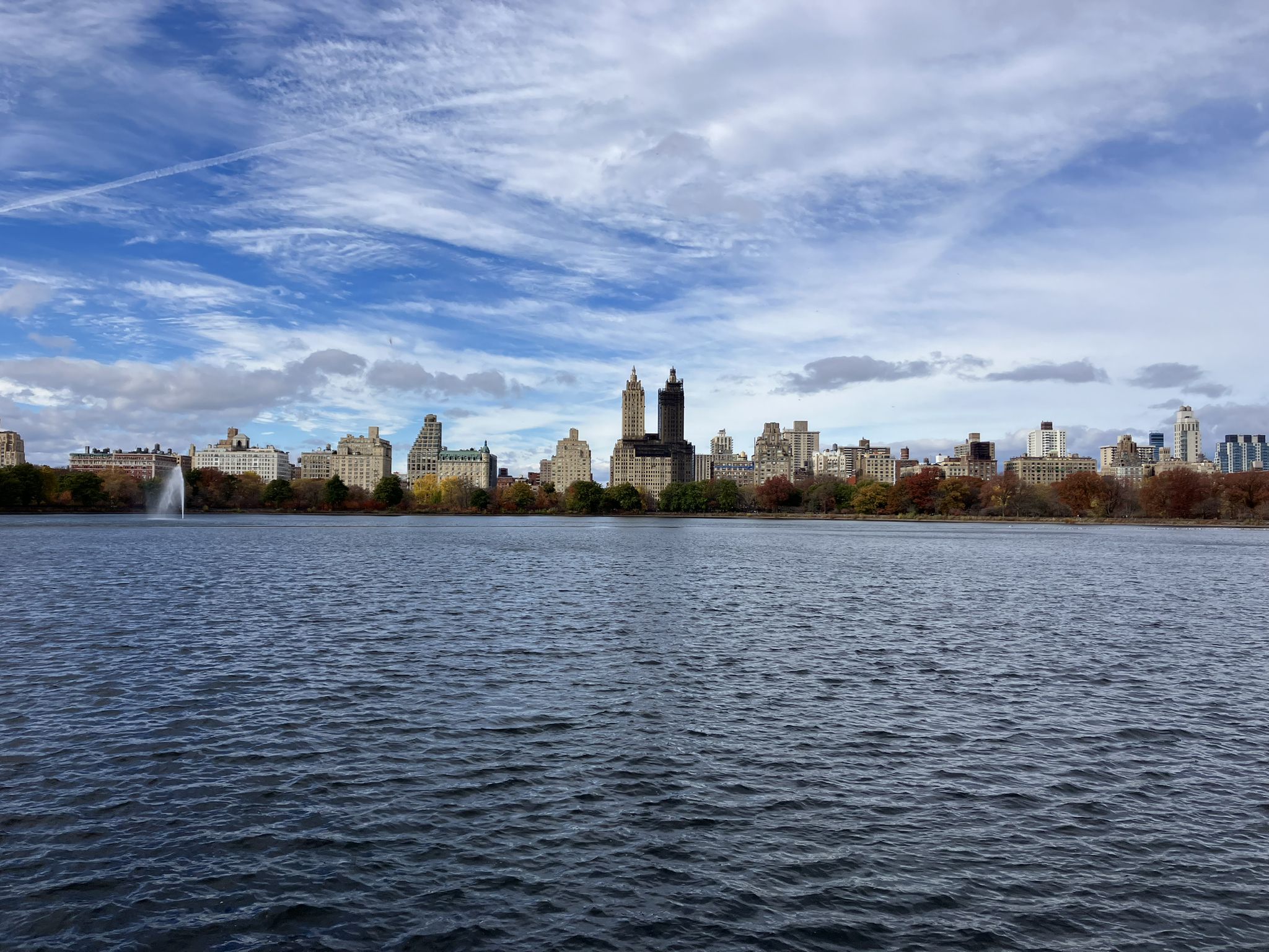 Lake with a distant city skyline and fountain under a cloudy sky. - Center Drive, Central Park – Harlem Meer loop from 125th Street