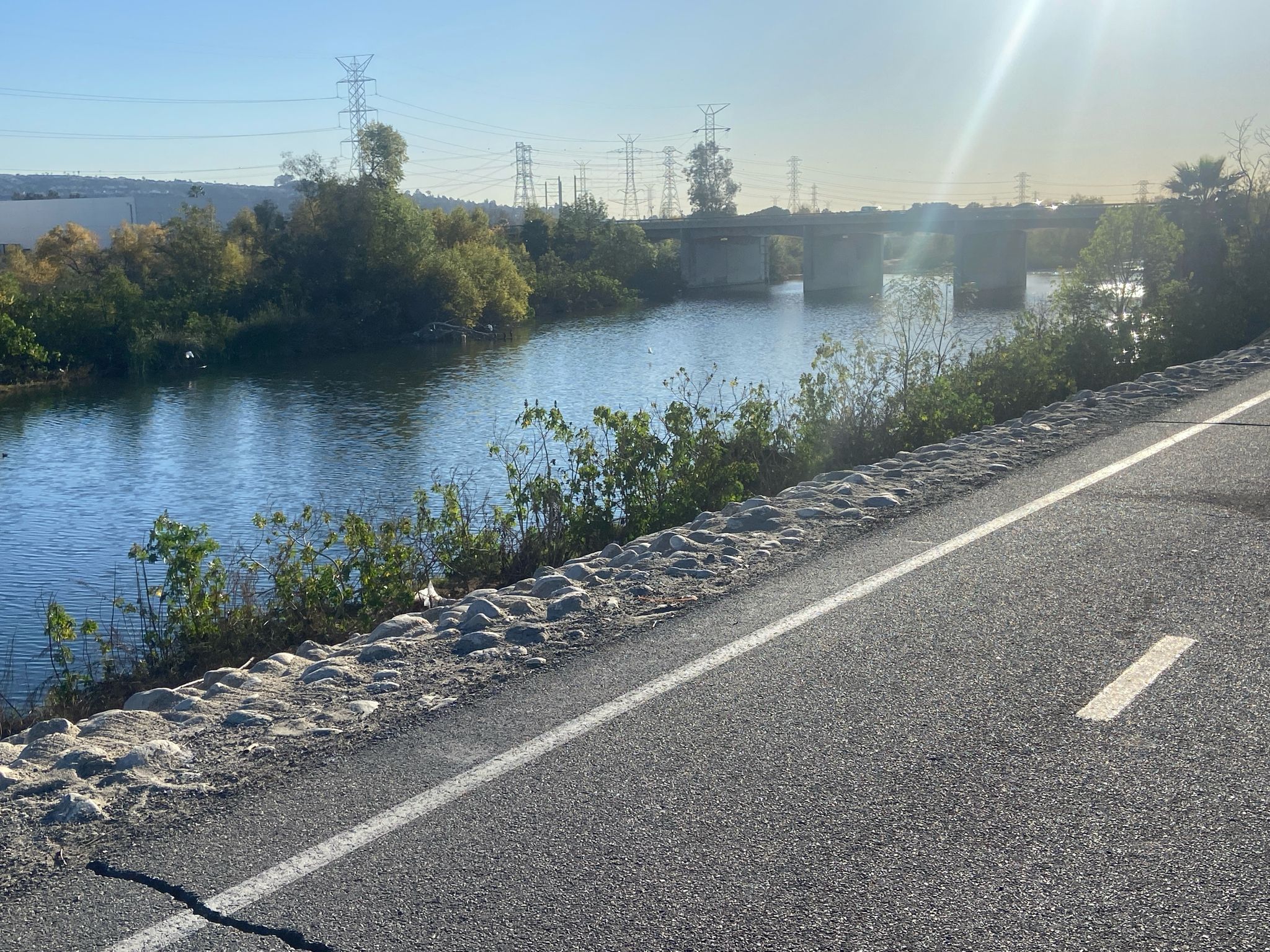 A paved path runs beside a river with a bridge and power lines. - Bosque del Rio Hondo – San Gabriel River Dam loop from Stonewood Center