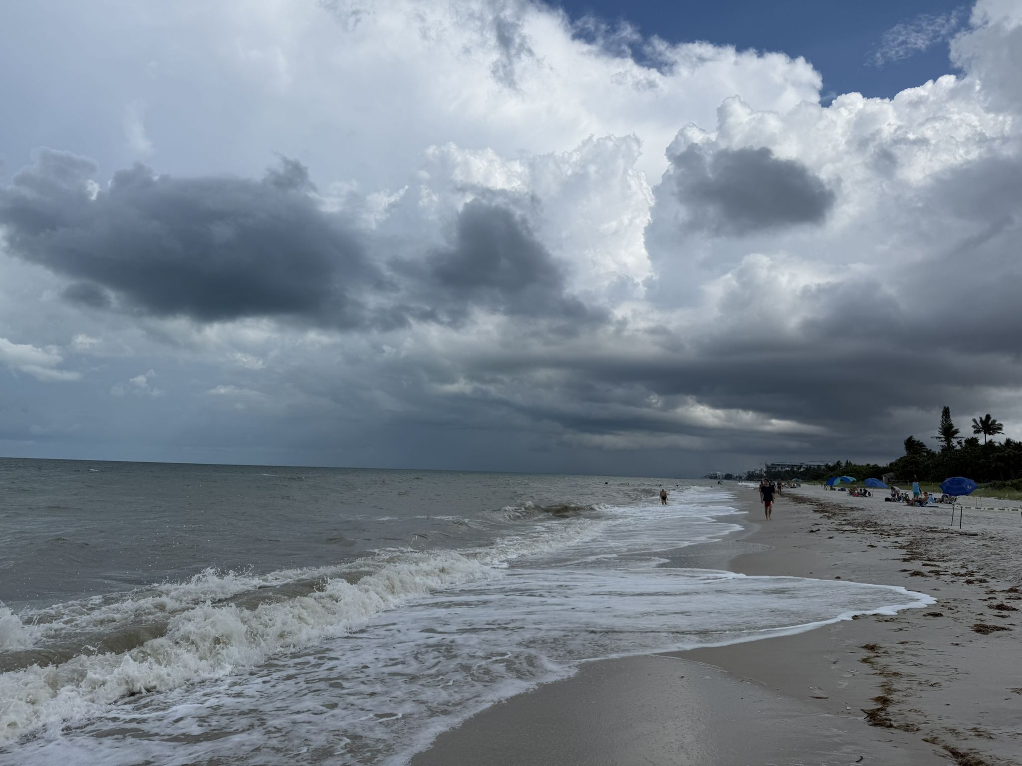 A stormy beach scene with dark clouds, crashing waves, and distant people. - Naples Beach via Lowdermilk Park