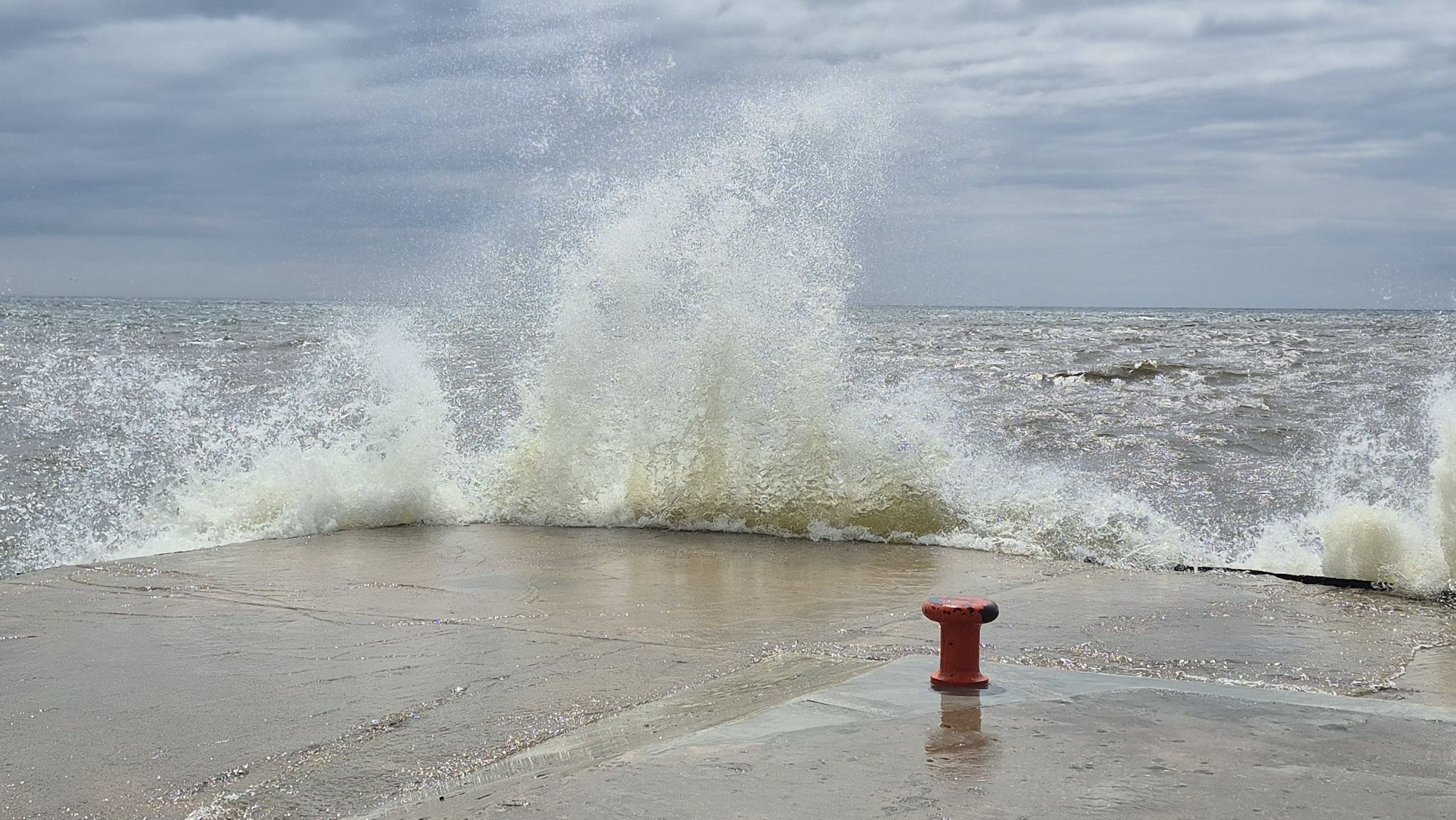 Large wave crashes over a concrete pier with a red bollard under an overcast sky. - Grand Haven Boardwalk