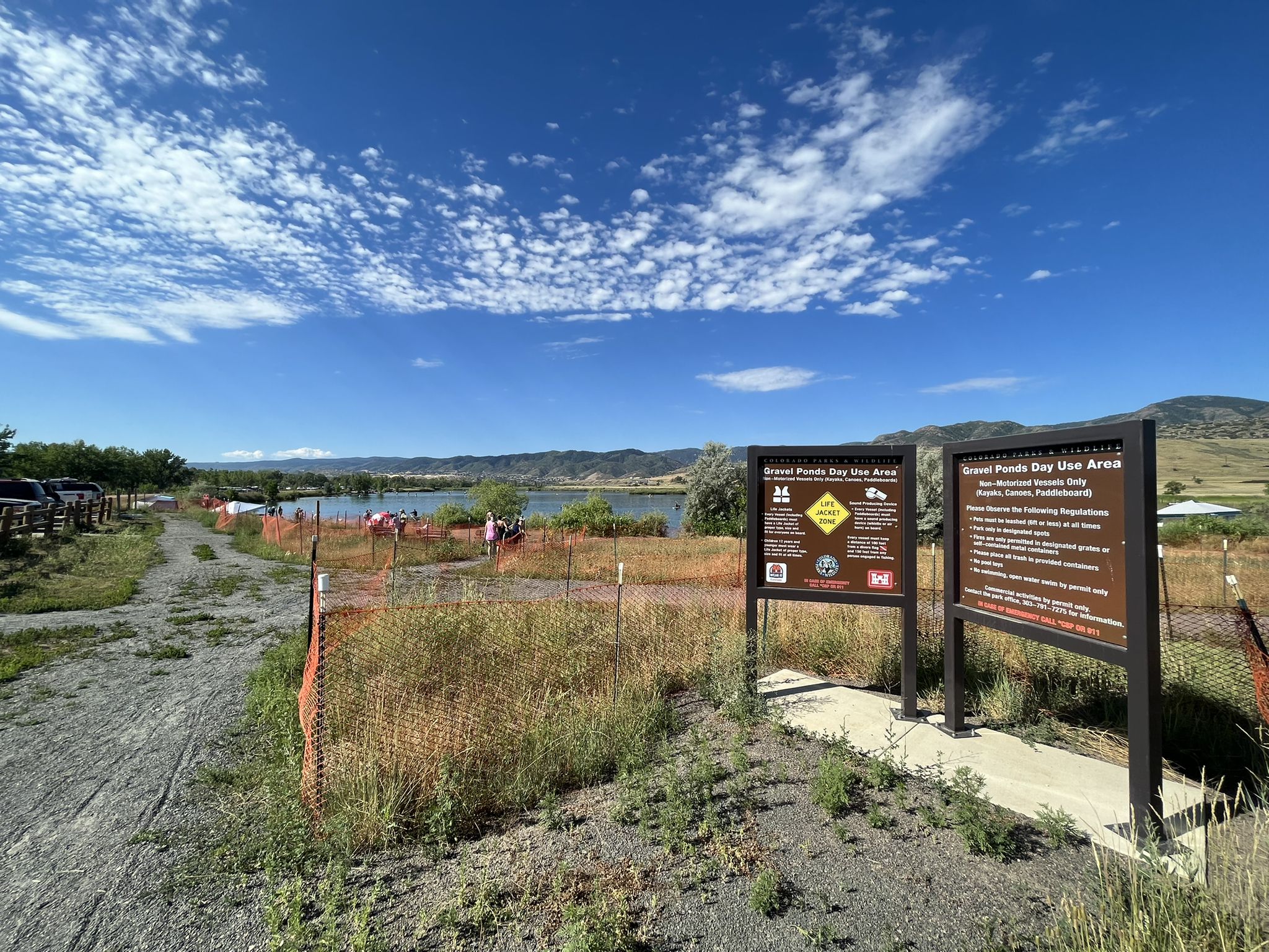 Two large signs beside a dirt path leading to a lake and mountains under a bright sky. - Wetlands View – Chatfield State Park loop from Kassler trailhead