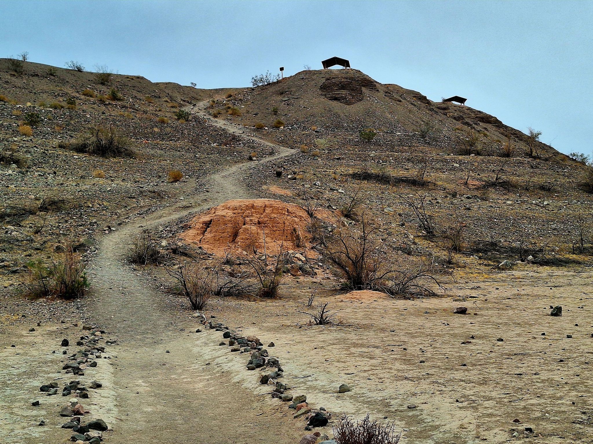 Winding dirt trail ascends a rocky, arid hill with small structures. - White Owl Canyon Trail