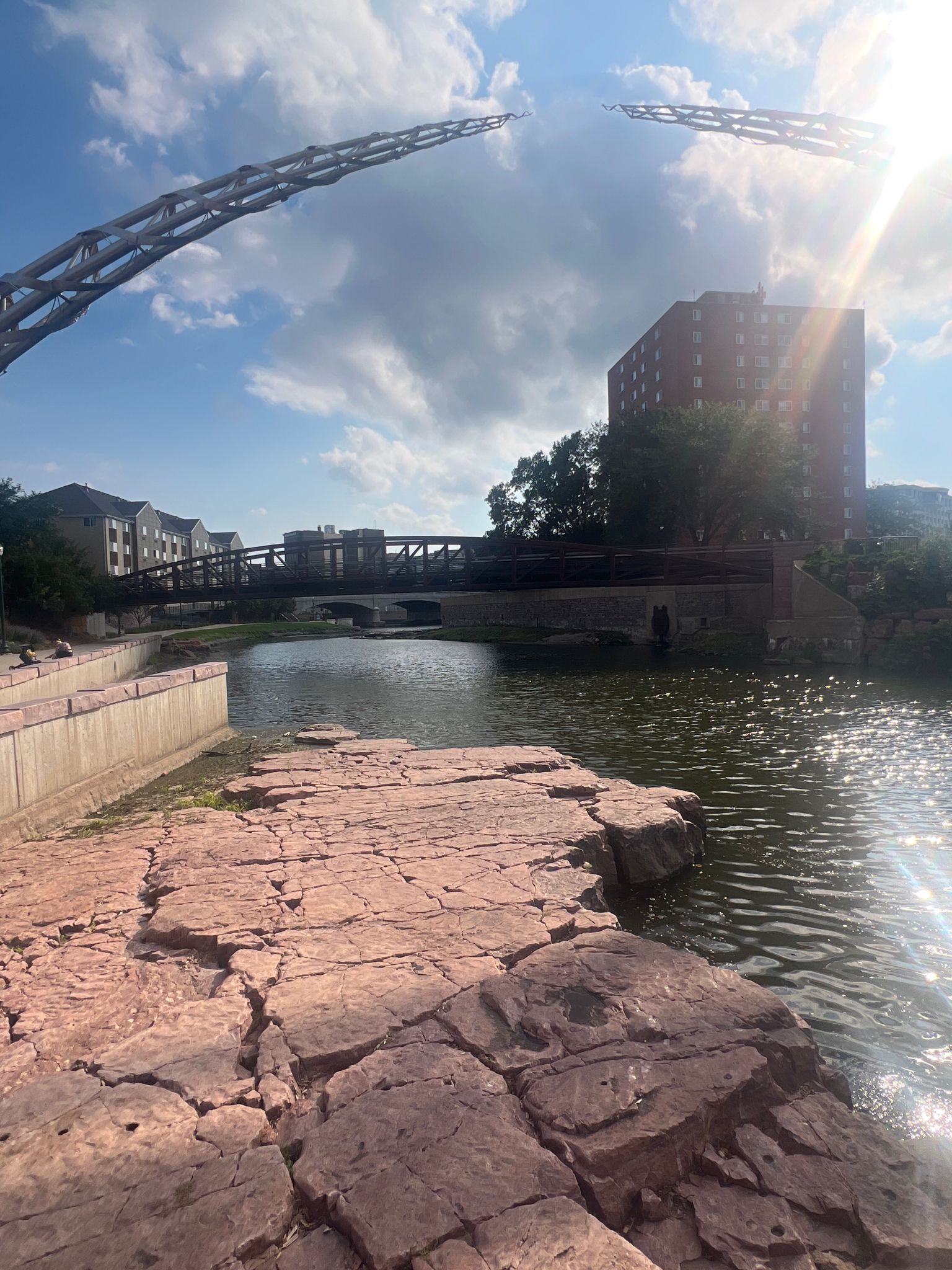 River with large red rocks, urban buildings, and bridges under bright sun. - Viewpoint (east side) – The Arc of Dreams loop from Sioux Falls