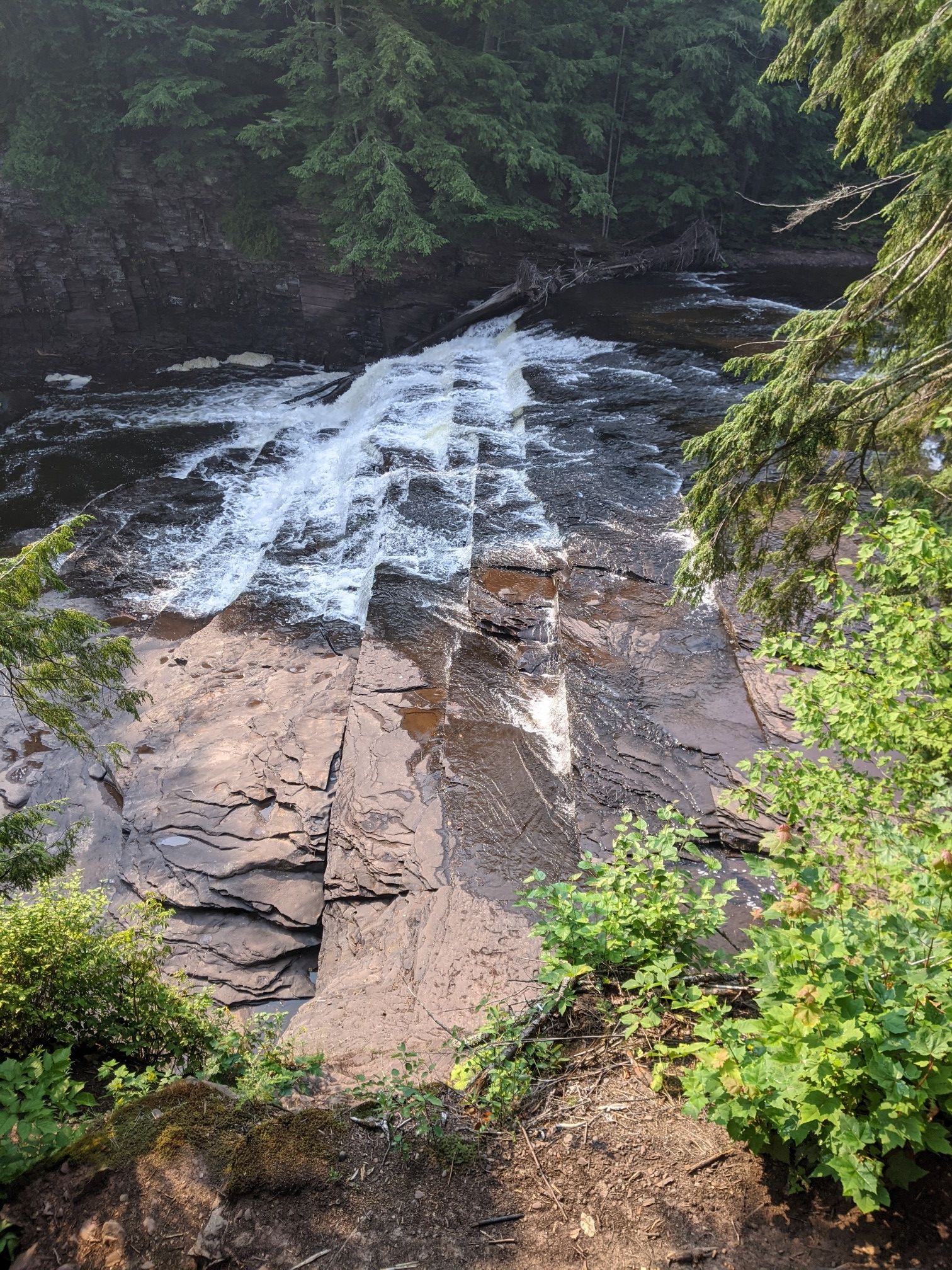 Waterfall cascades over dark layered rocks, surrounded by dense green forest. - Presque Isle River Waterfalls Trail