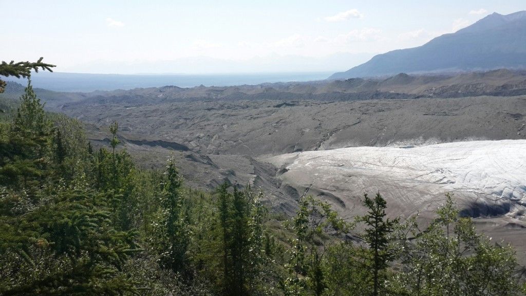 Overhead view of a glacier and rocky terrain with green trees and hazy sky. - View of Root Glacier – View of Root Glacier loop from Glacier View 2025