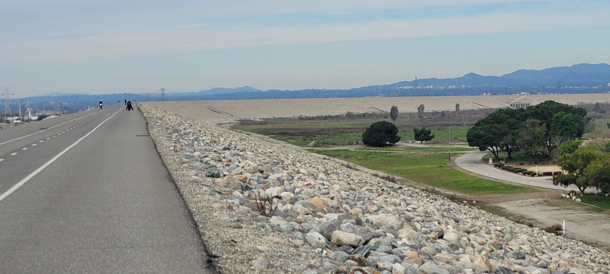 A paved path along a rocky embankment with cyclists and green parkland. - Santa Fe Flood Control Basin loop from Irwindale