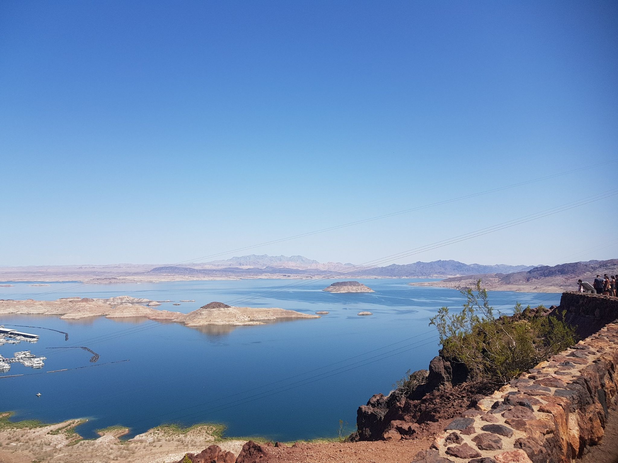 Vast blue lake with islands, distant mountains, and a clear sky. People on an overlook. - River Mountains Loop Trail
