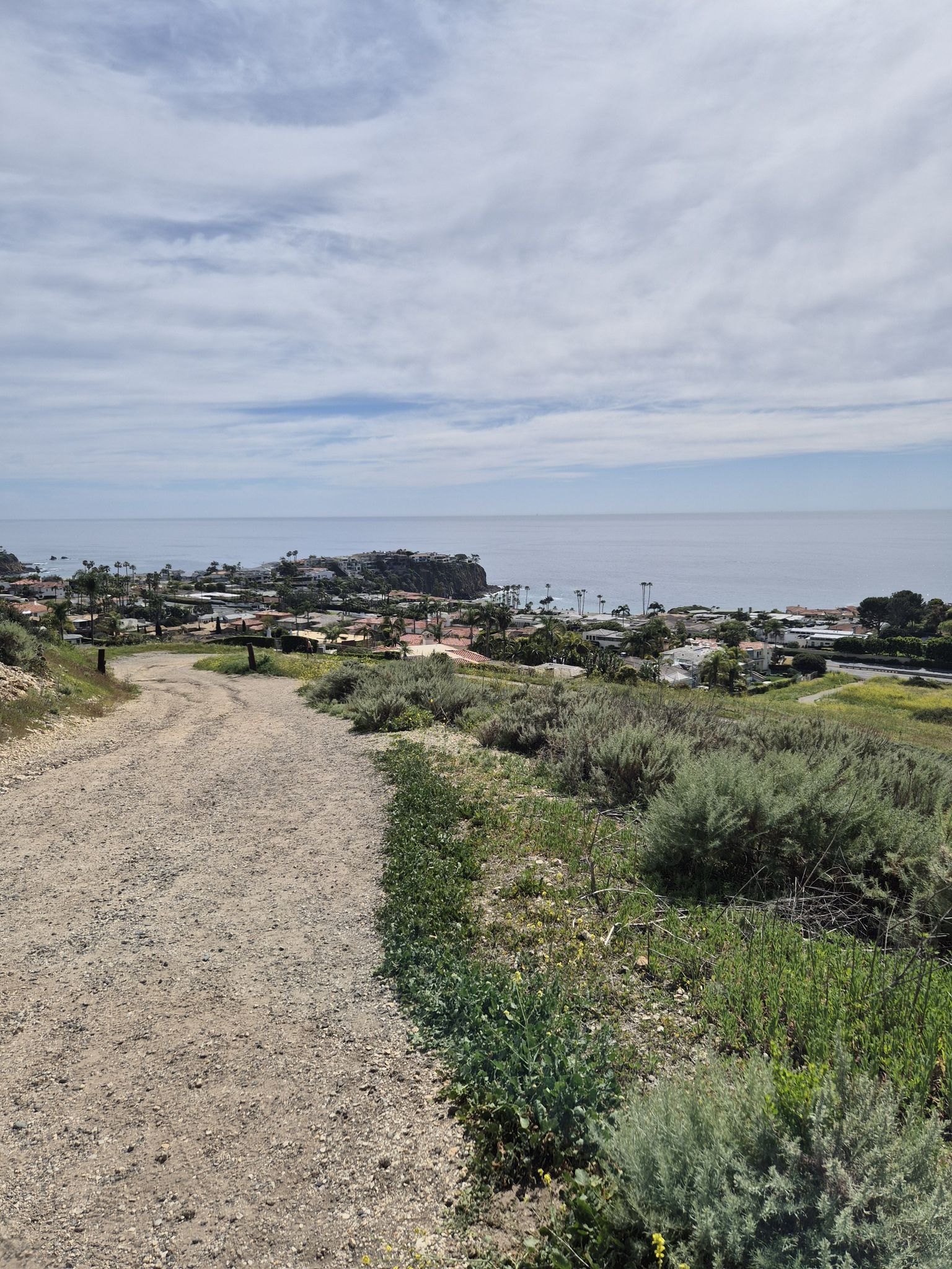 A dirt path descends towards a coastal town and the ocean under a partly cloudy sky. - El Moro Canyon to Moro Ridge Loop