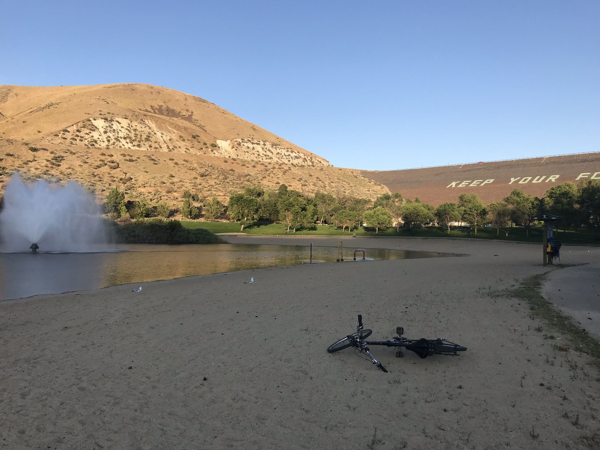 A bicycle on a sandy beach next to a lake with a fountain and distant hills. - Boise River Greenbelt