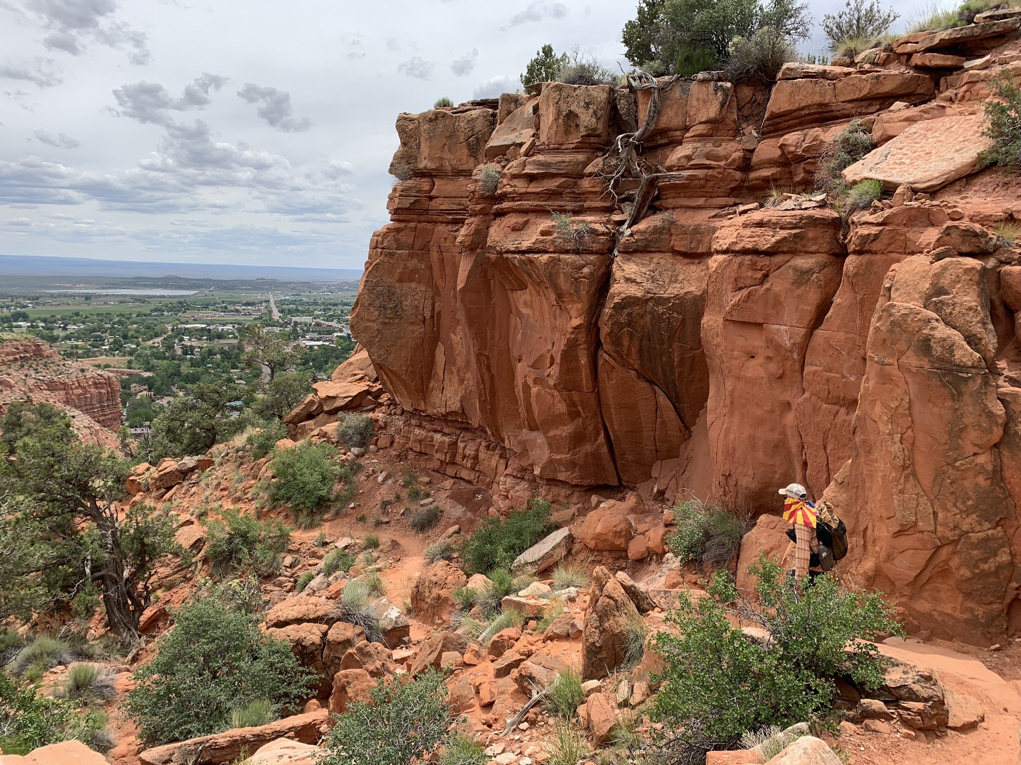 Hiker stands on a trail beside massive red rock cliffs, overlooking a town. - Kanab View Boulder loop from Kanab