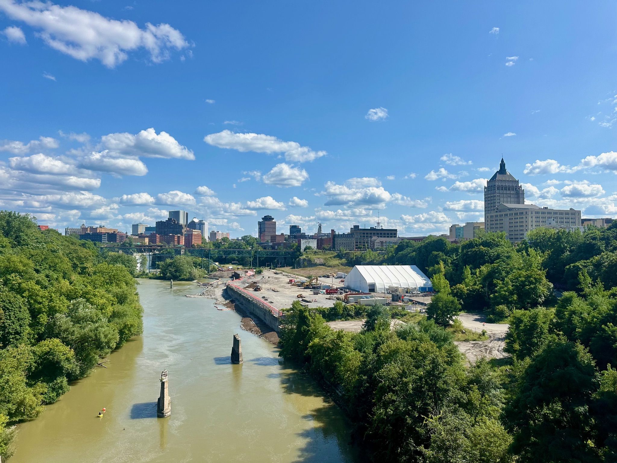 River flows past a city skyline and construction under a bright, cloudy sky. - Genesee Riverway Trail – High Falls loop from Rochester