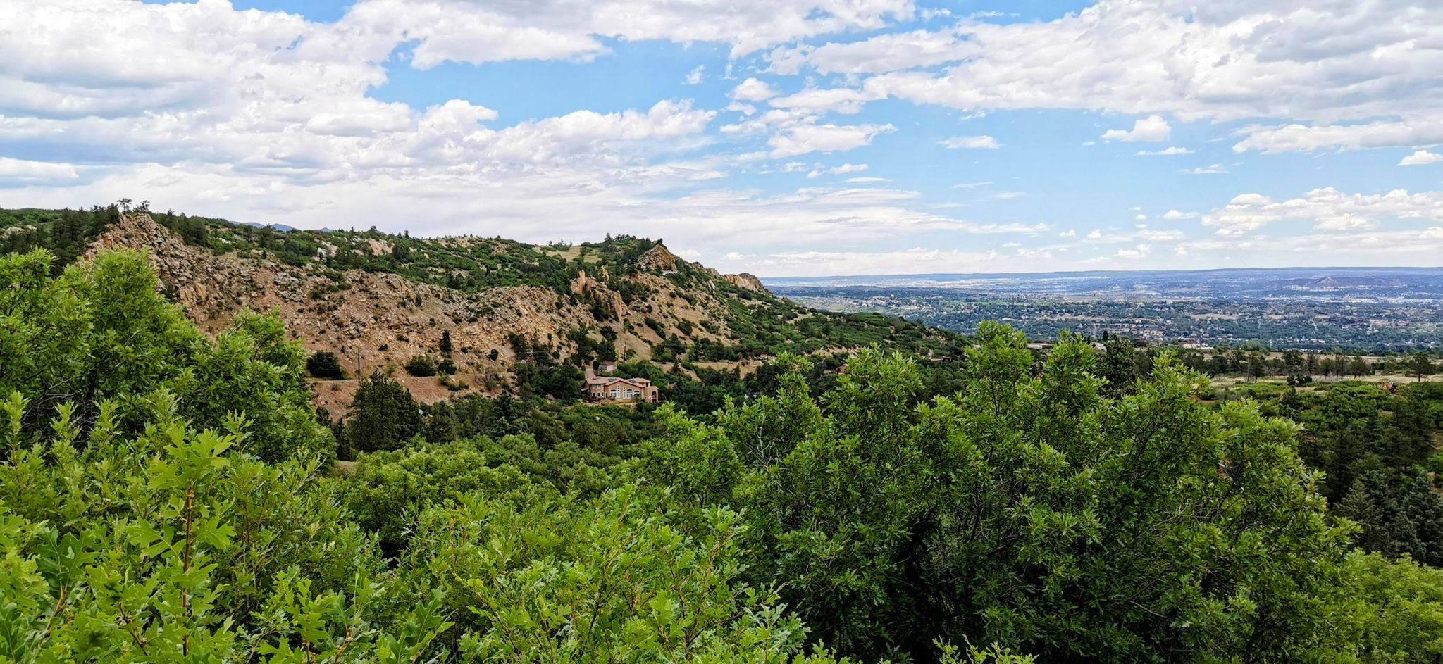 Lush green trees and rocky hills overlook a distant city under a cloudy sky. - Cripple Creek from Colorado Springs on the Gold Camp Road
