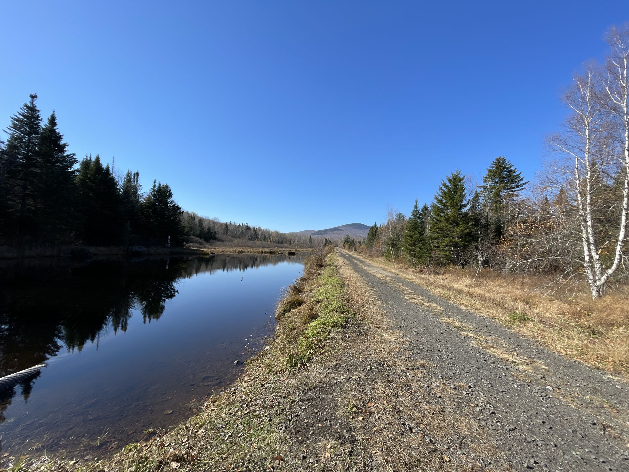 Gravel path next to a calm lake, distant mountains, clear sky. - Cross New Hampshire Adventure Trail and Pine Mountain Loop