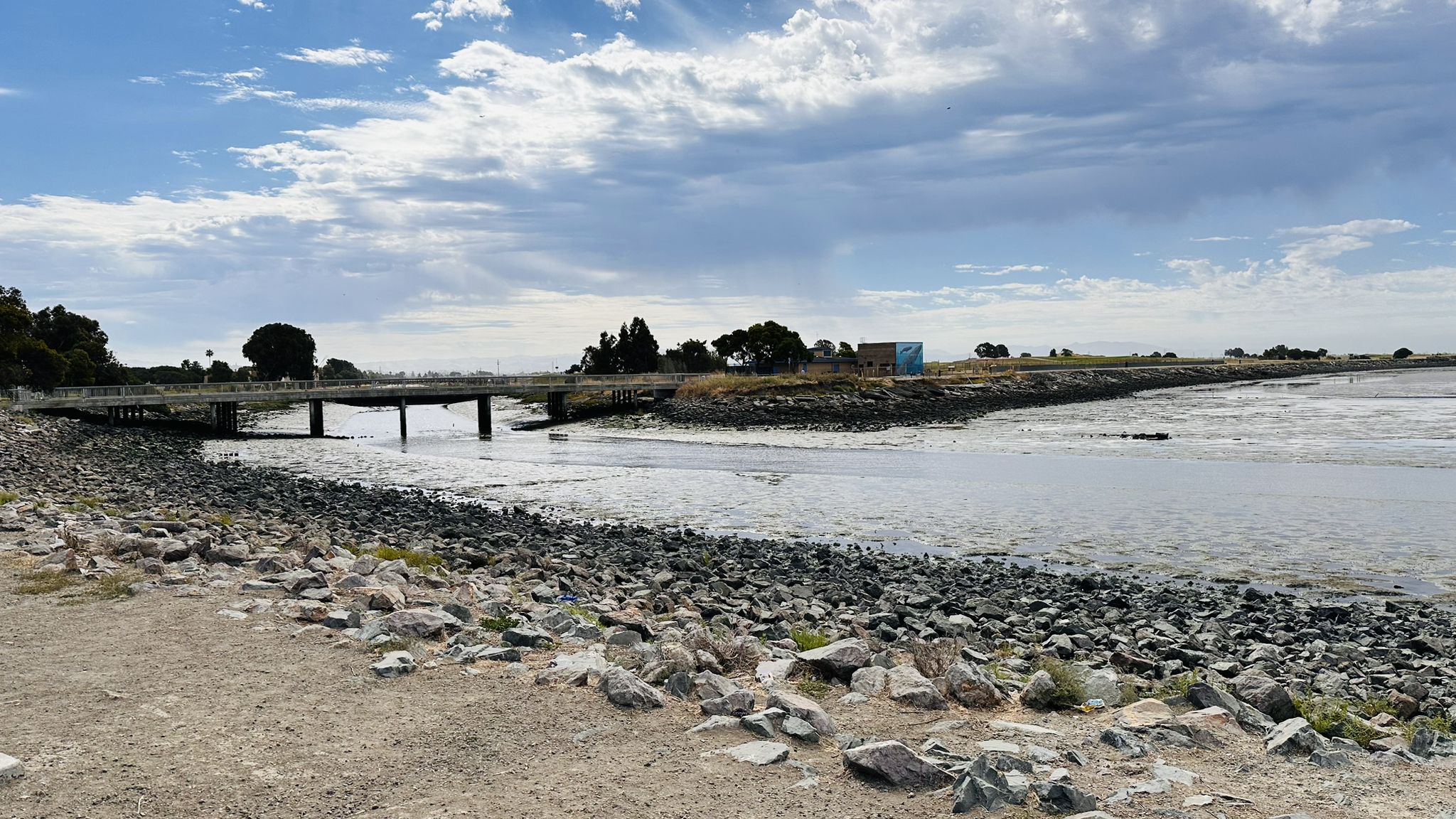 Rocky shoreline and calm water with a distant bridge under a cloudy sky. - Marina Park, San Leandro – Wes McClure Boat Launch loop from Oyster Bay Regional Shoreline