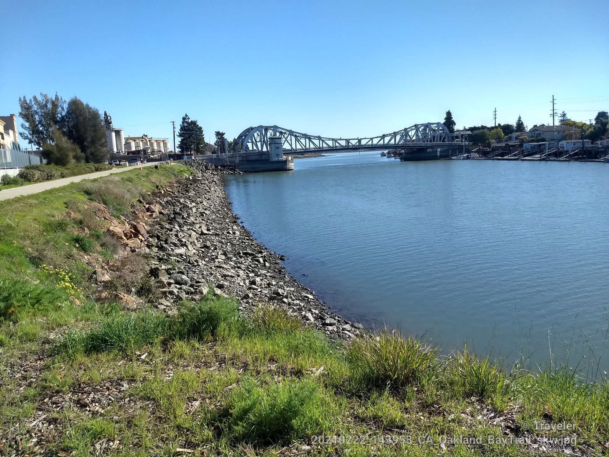 A river with a large bridge, rocky shore, and green path under clear sky. - San Francisco Bay Trail: Alameda Loop