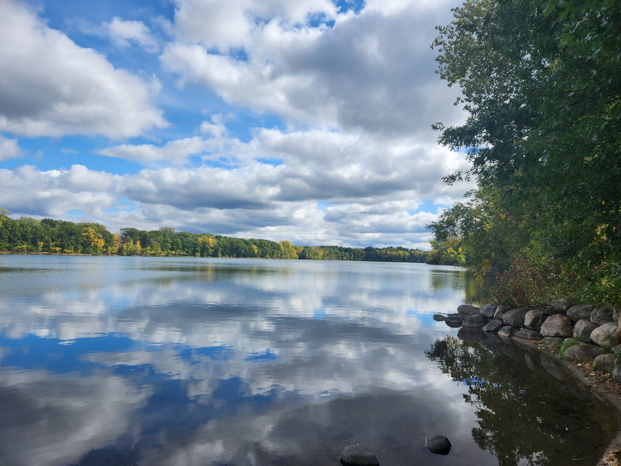 Calm lake reflecting cloudy sky, surrounded by green trees and rocky shore. - Powers Lake Trail