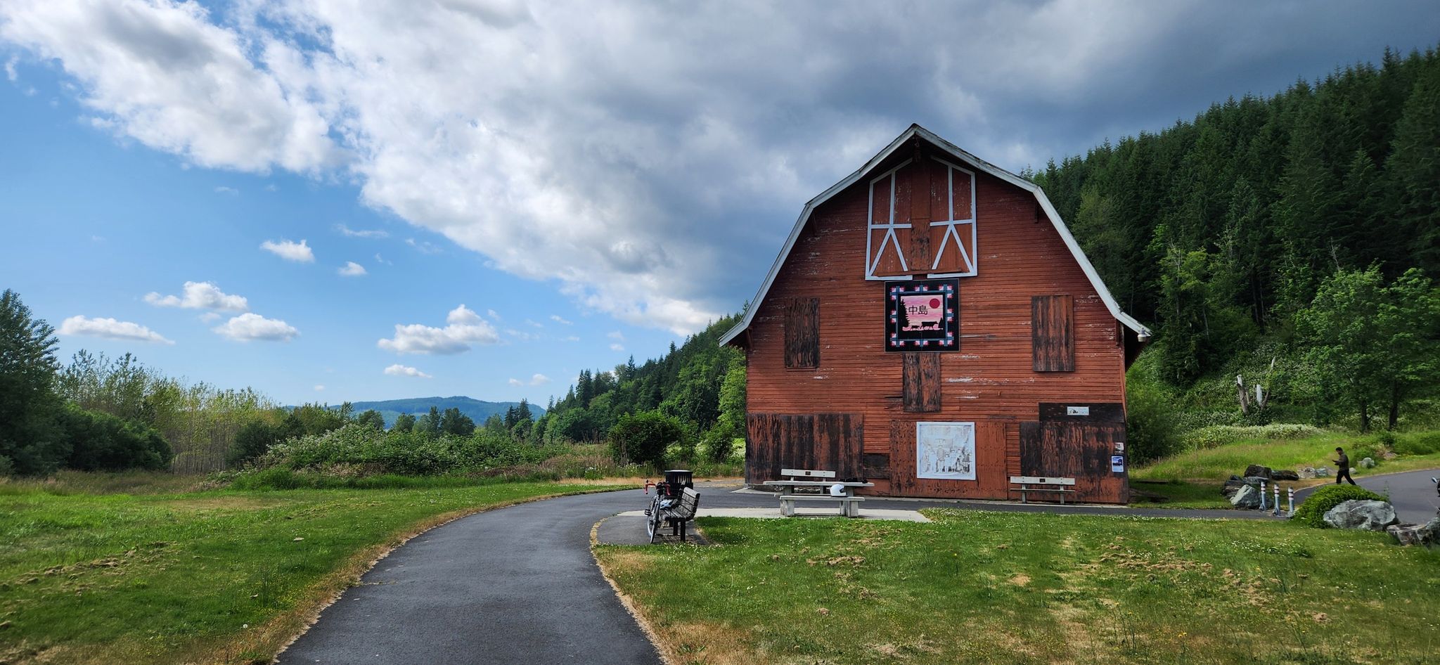Red barn by a paved path, forested hills, and cloudy sky. - Bryant to Lake McMurray via Centennial Trail