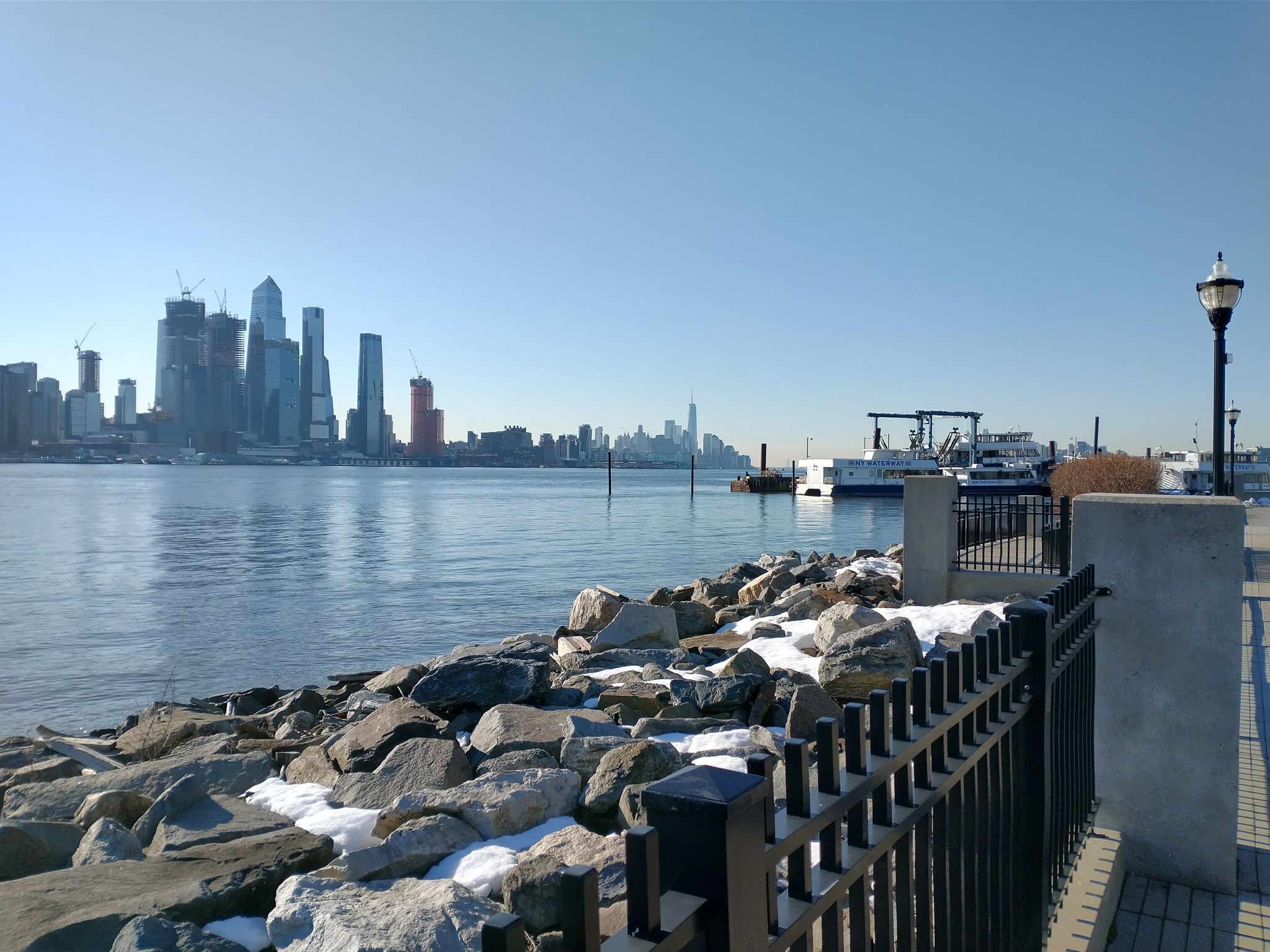 Rocky shore with snow patches, city skyline across water on a sunny day. - Port Imperial Waterfront loop from Columbia University, Manhattanville Campus