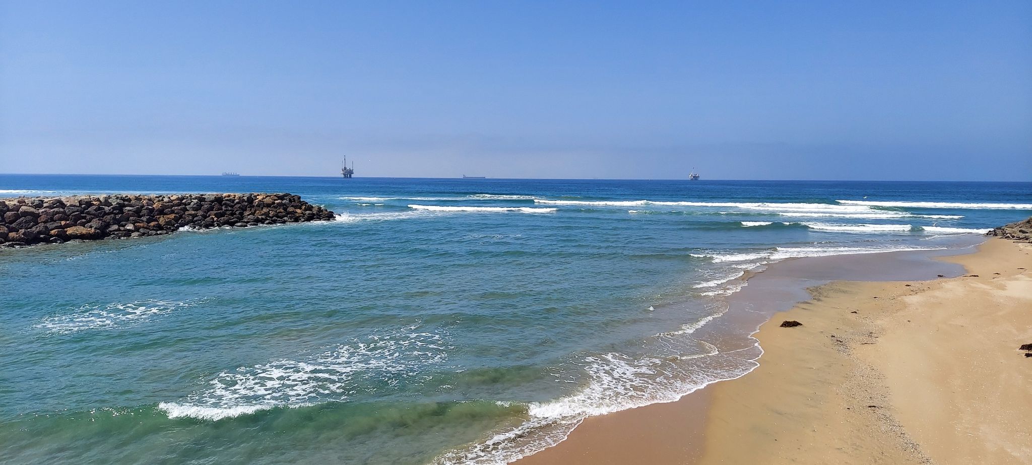Sandy beach with ocean waves breaking near rock jetties on a sunny day. - Bolsa Chica Ecological Reserve Trail