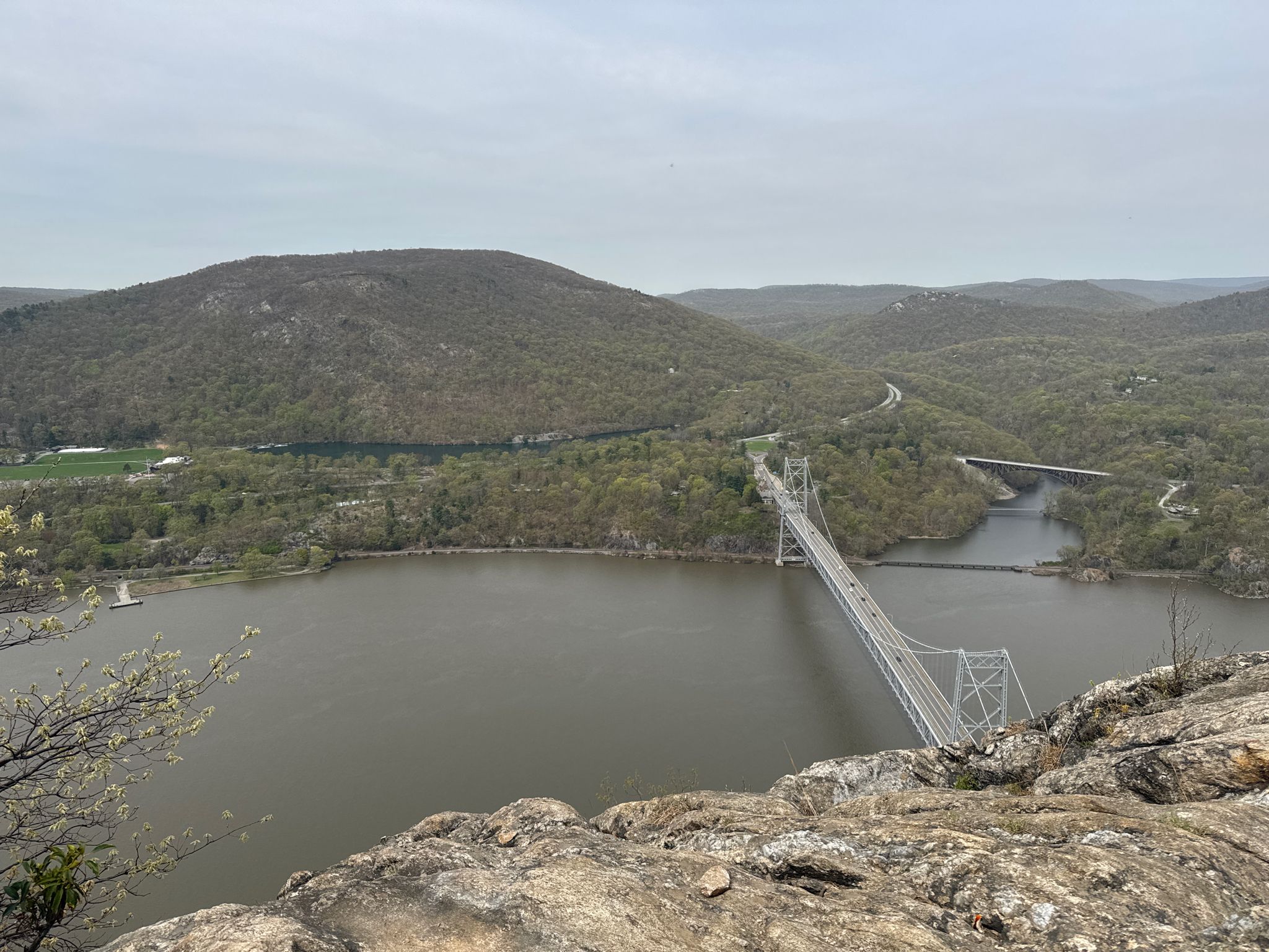 Overcast view of a wide river, forested hills, and a large bridge. - Anthony's Nose via Camp Smith Trail