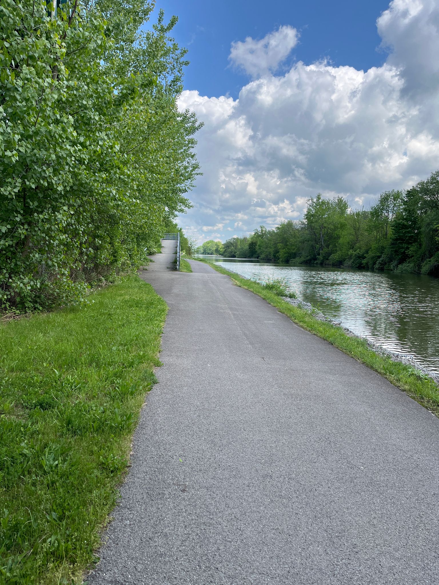 A paved path runs beside a river with green trees on a bright cloudy day. - Bridge over the Erie Canal loop from Perinton