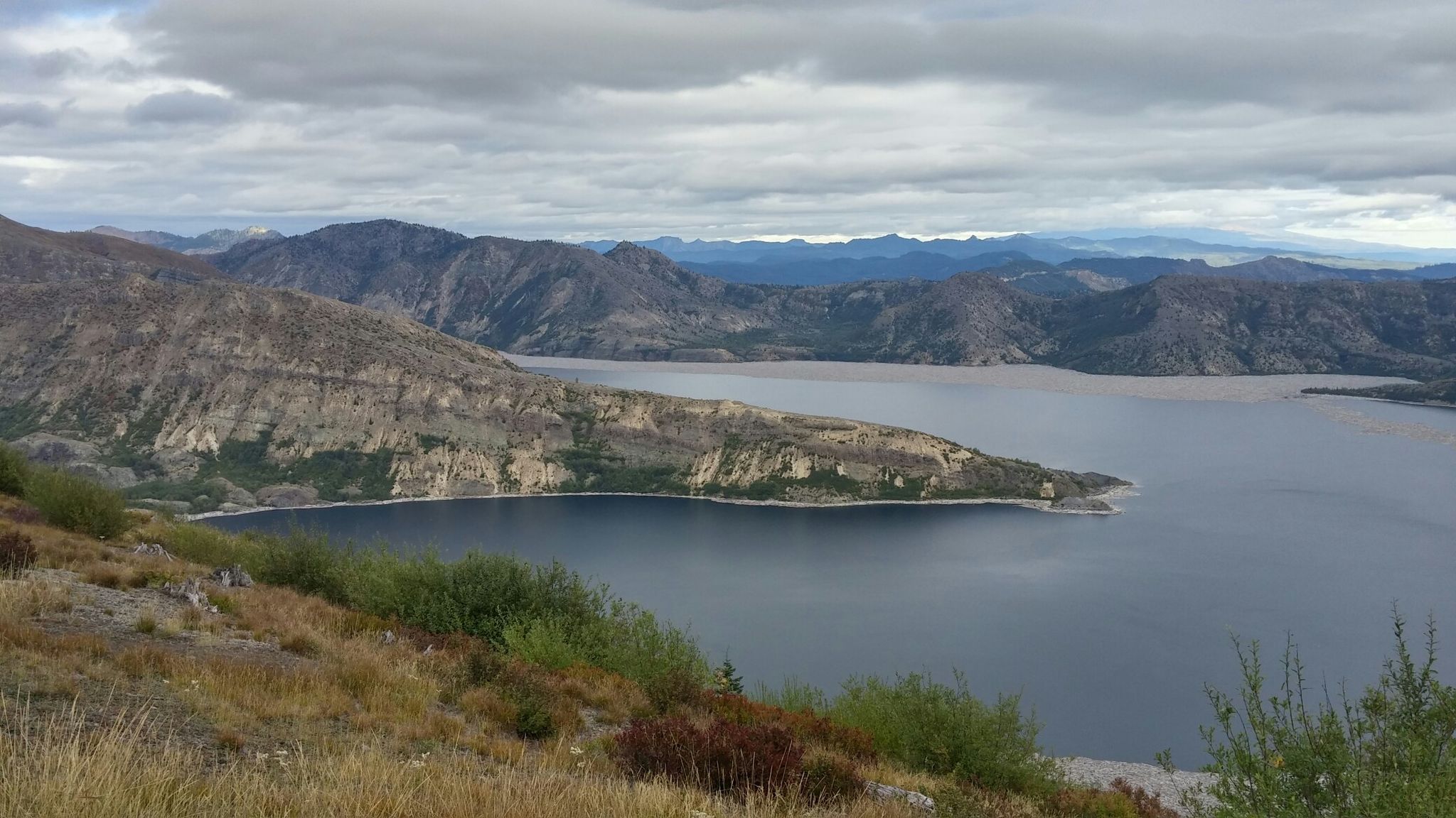 Overcast panoramic view of a wide lake and surrounding rocky, green mountains. - Harry's Ridge Trail