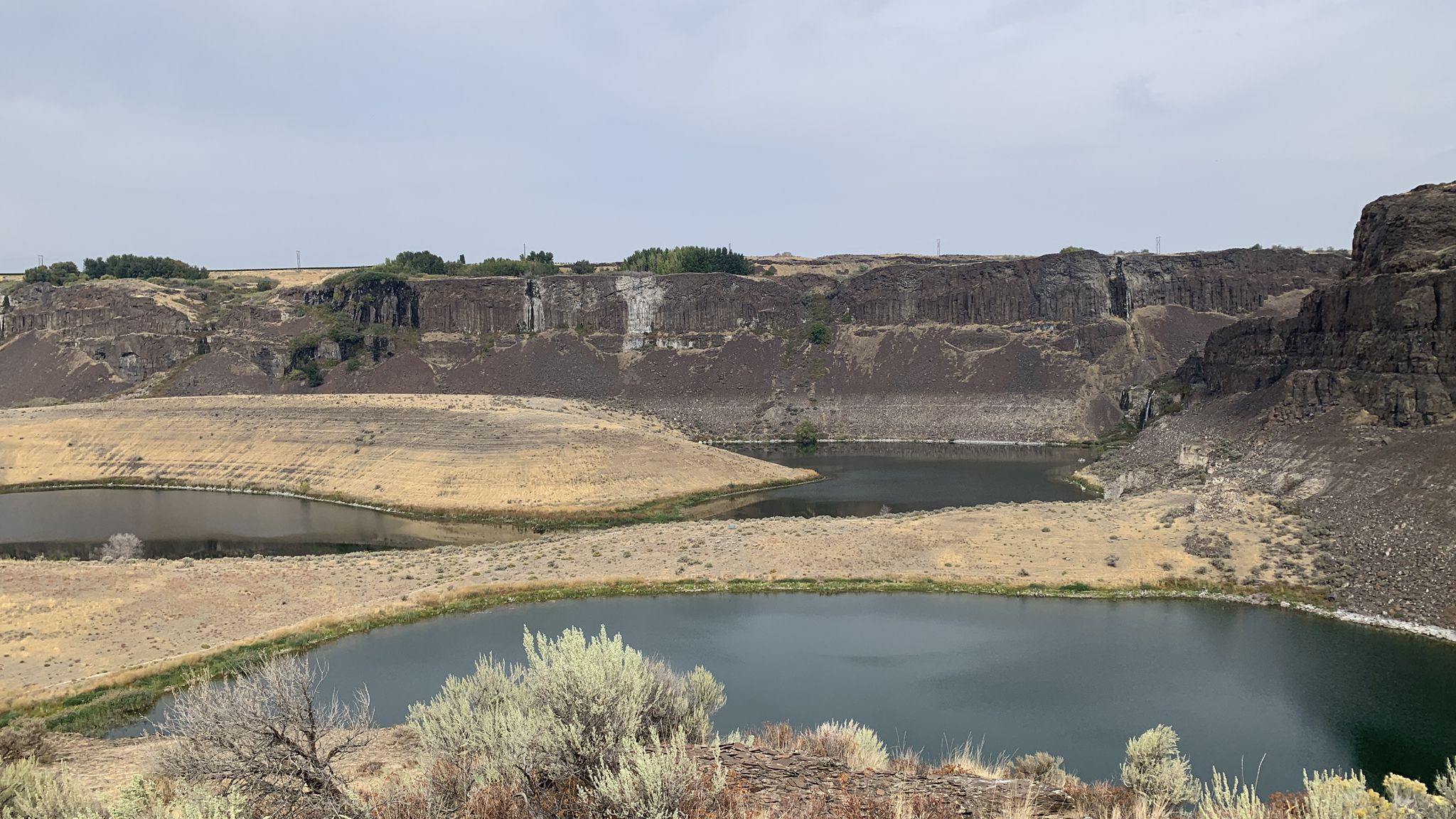 Two dark lakes nestled among dry hills and rugged cliffs under an overcast sky. - Dusty Lake Trail
